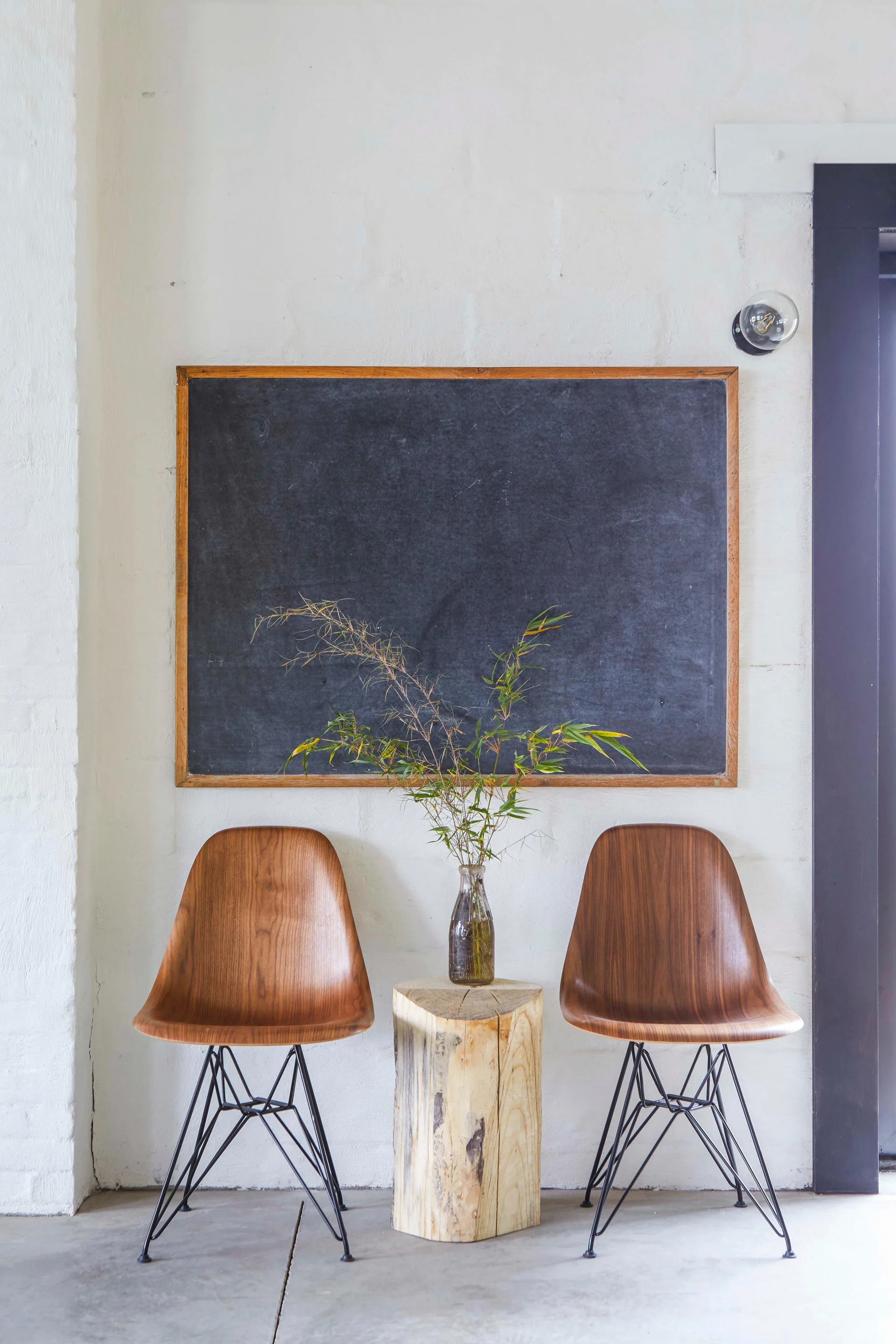 Two molded wood chairs with black metal legs flank a natural log stool beneath a framed chalkboard on a painted white brick wall.