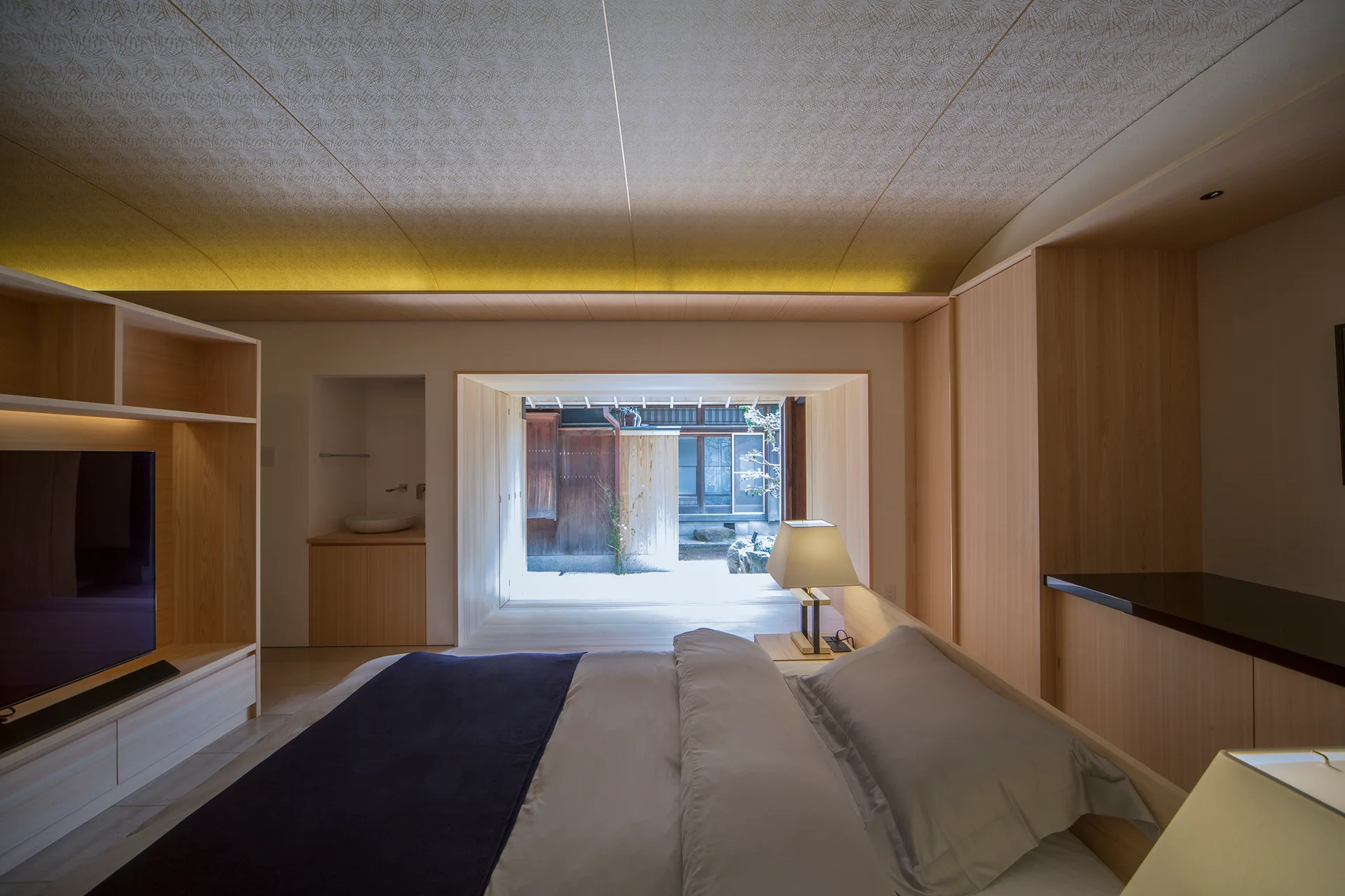 A minimalist bedroom features light wood built-in cabinets, a bed with grey linens, and a large open doorway looking out to a bright courtyard.