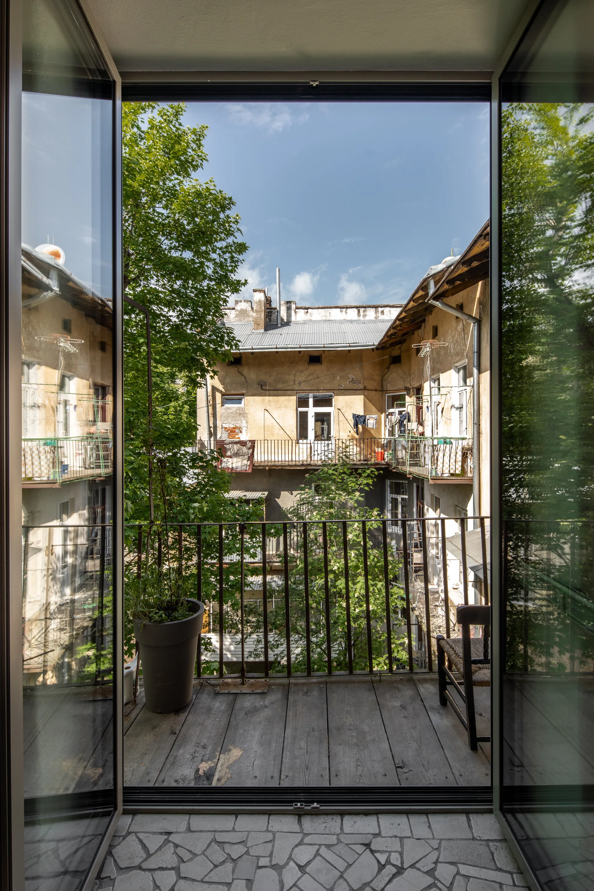 Open glass doors look out onto a narrow wooden balcony with a metal railing and a large potted plant, overlooking a central urban courtyard.