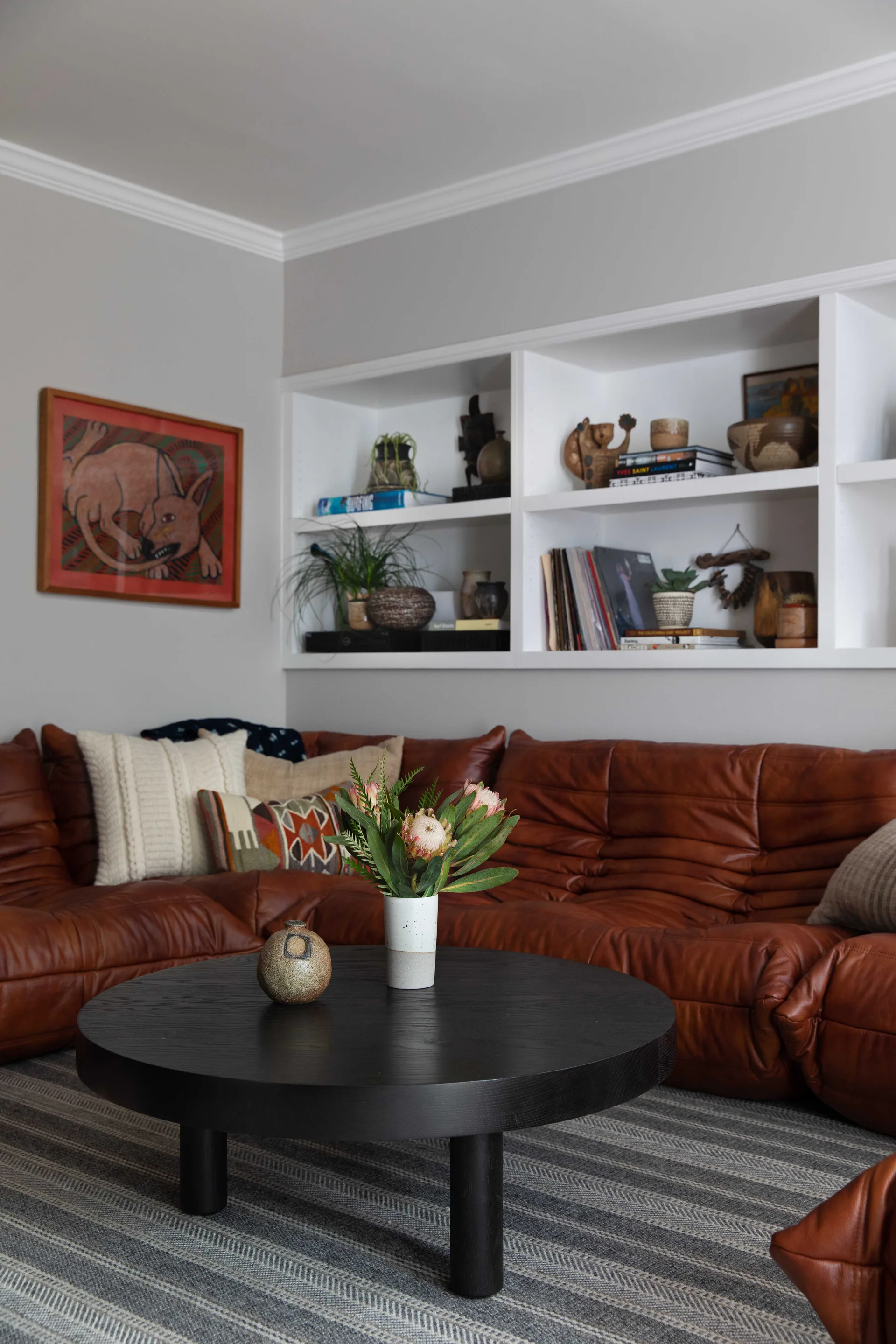 A living room features a low-profile brown leather sectional sofa around a round black wood coffee table, set against grey walls with white built-in shelves.