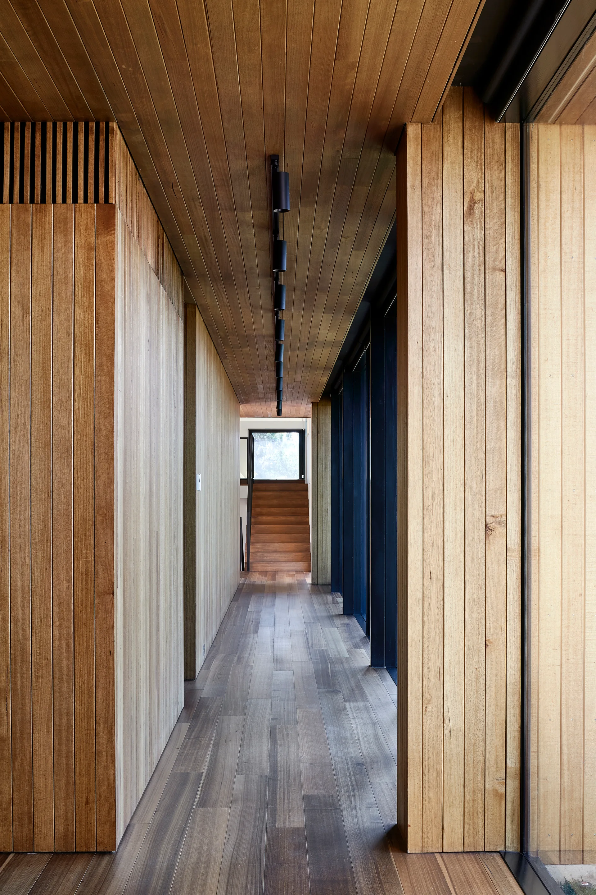 A long interior hallway clad entirely in vertical wood paneling features a matching wood ceiling, black track lighting, and a staircase at the end.