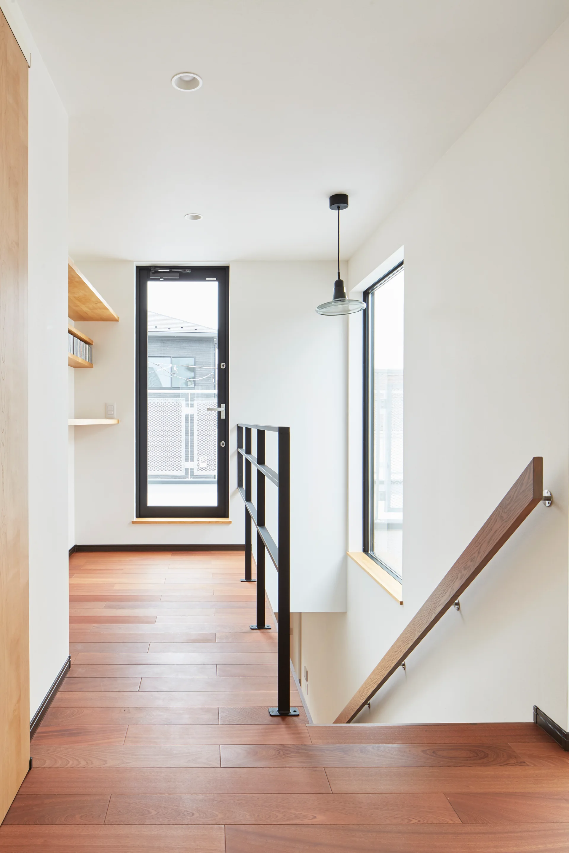 A bright modern staircase landing features warm wood flooring, a black metal railing, floating wooden shelves, and a black-framed glass door.