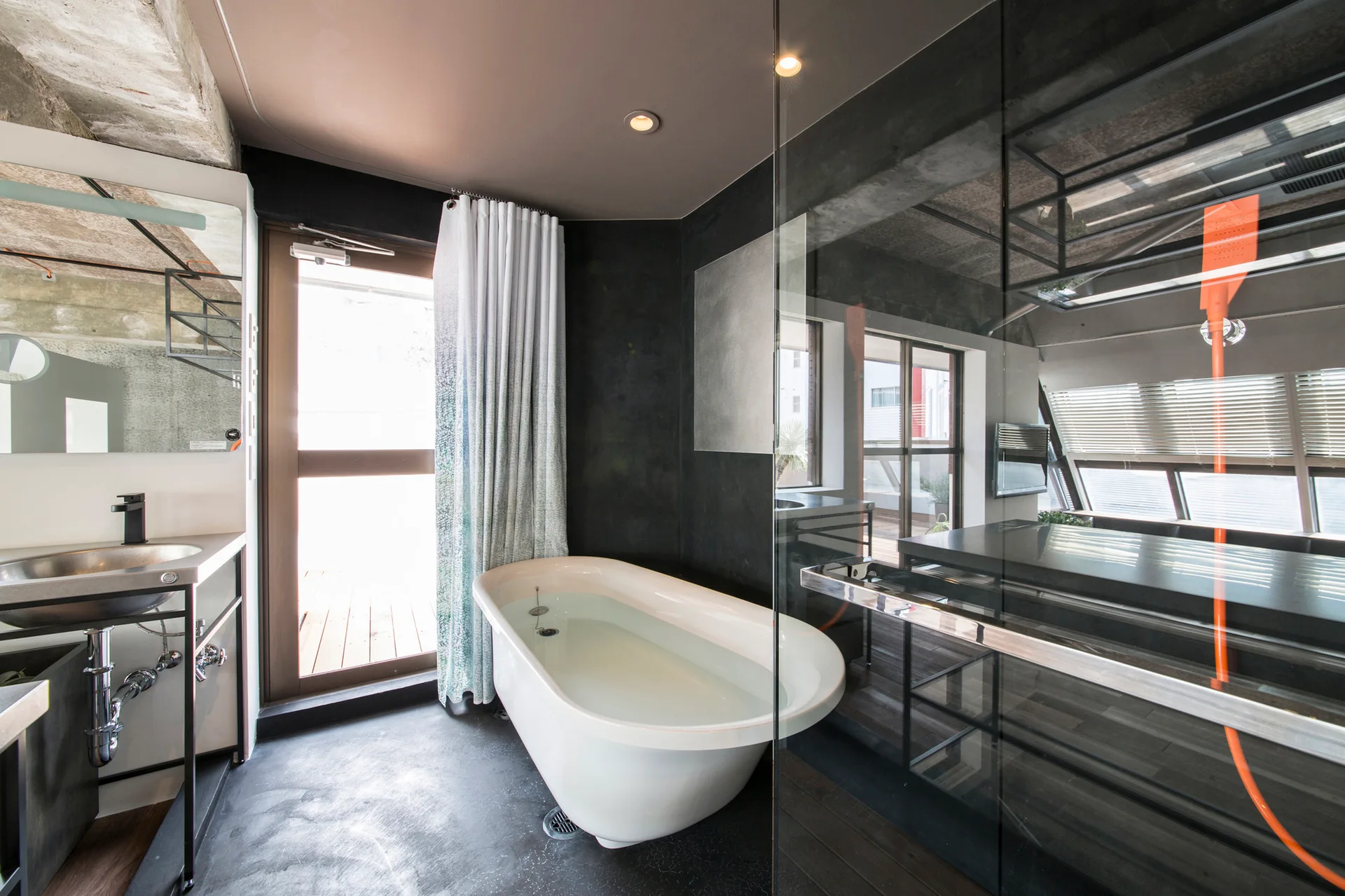 A modern industrial bathroom features a white freestanding bathtub next to a black wall, a metal sink console, and a large glass partition.