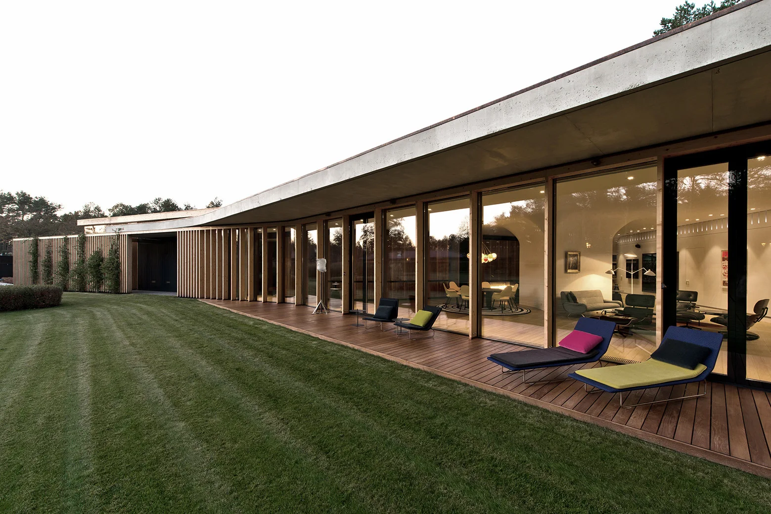 A modern curved home features a floor-to-ceiling glass facade, a wooden deck with outdoor chaise lounges, and an expansive manicured green lawn.