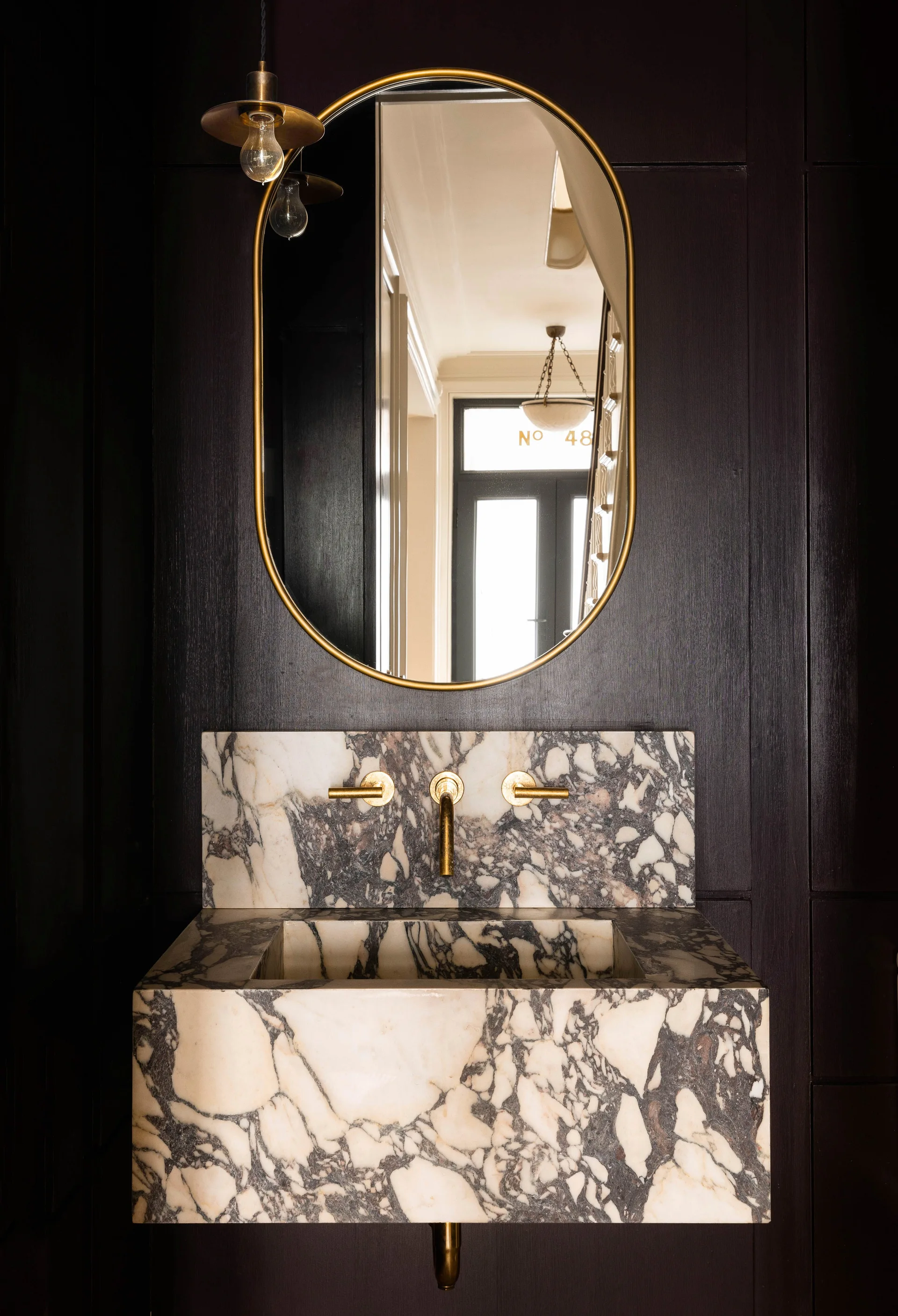 A wall-mounted marble sink with bold dark veining is paired with a brass faucet and an oval mirror against dark purple wood paneled walls.