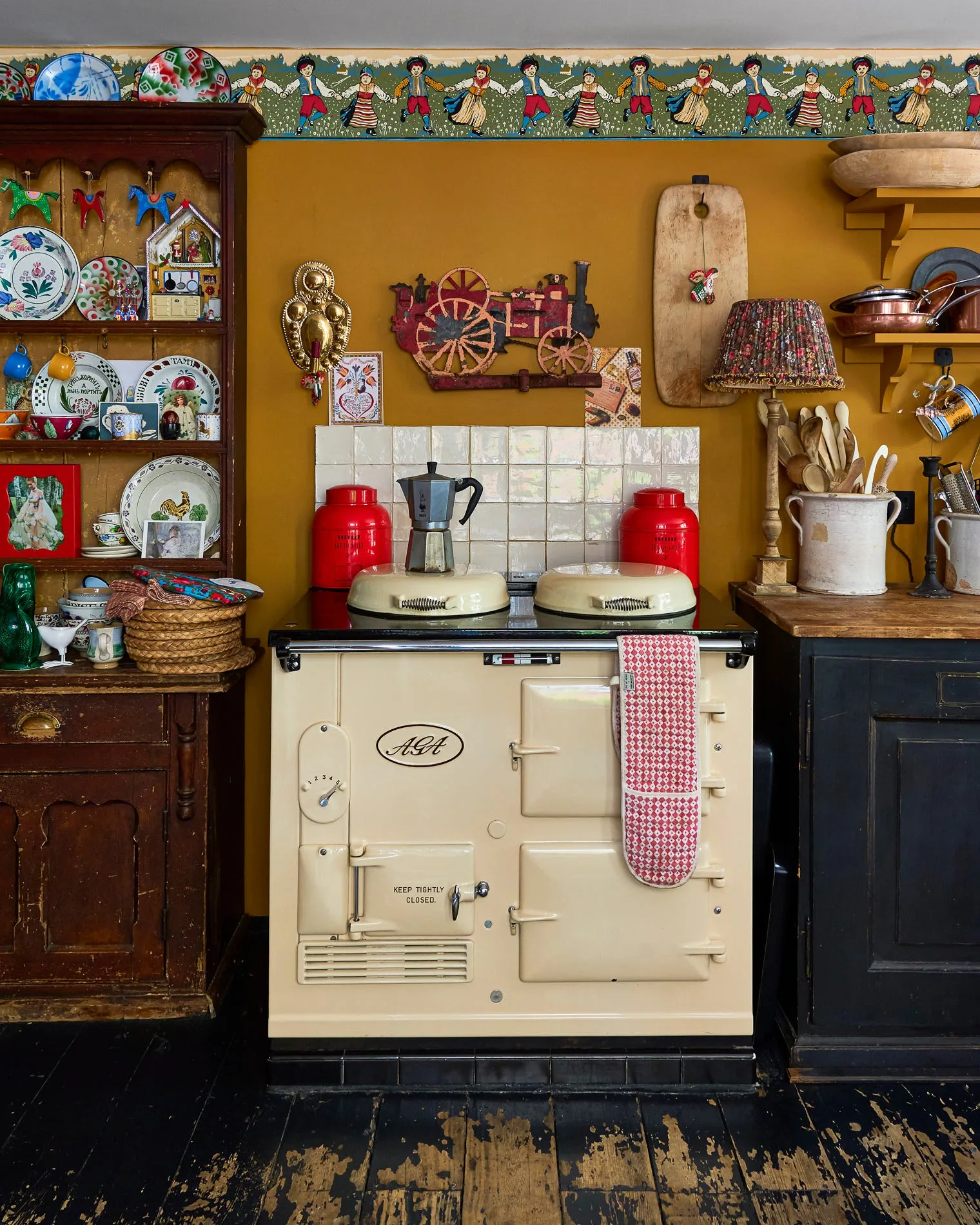 A cream-colored AGA stove sits against a mustard yellow wall decorated with a folk-art wallpaper border and flanked by a dark wood hutch and cabinets.