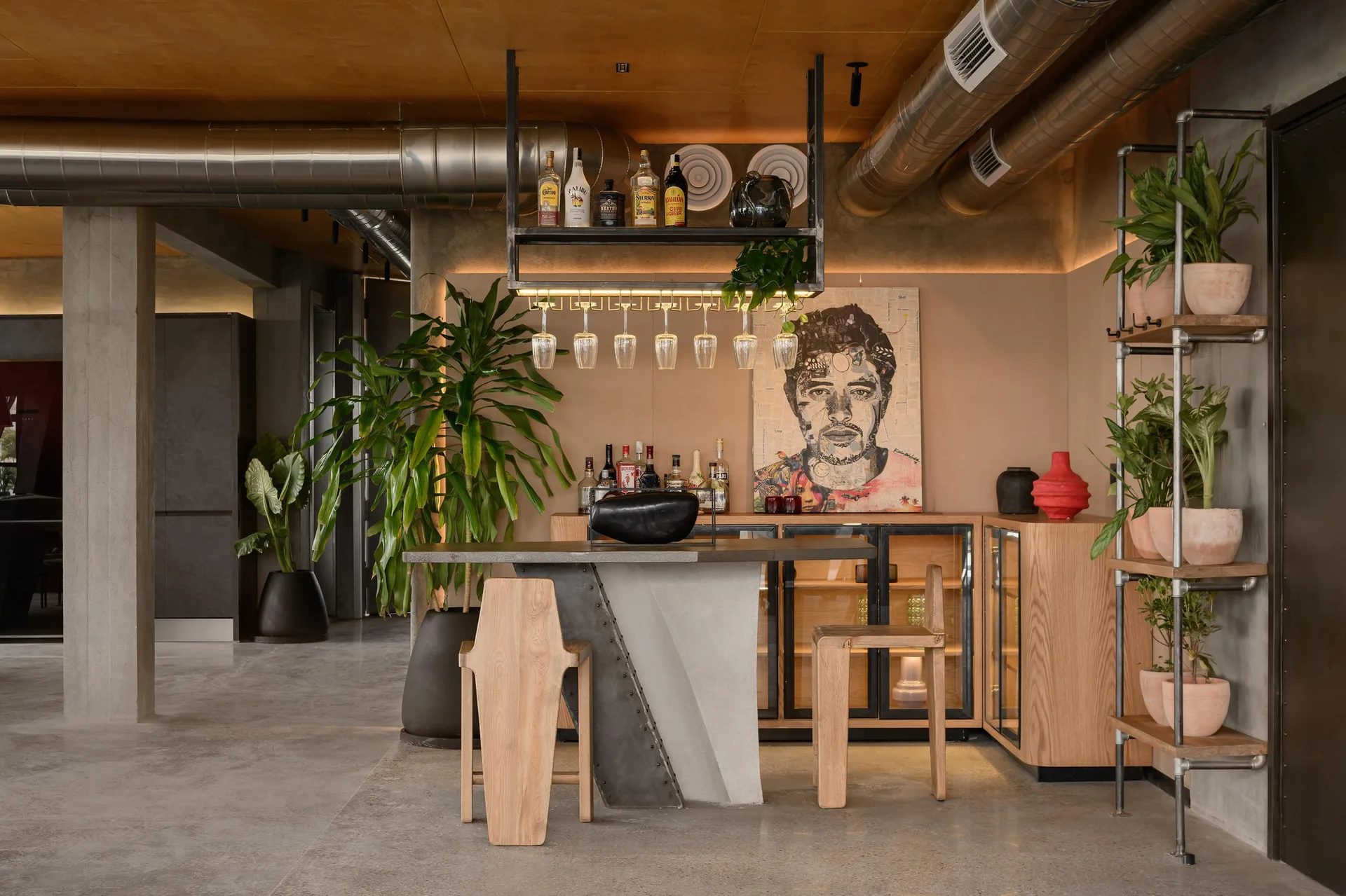 An industrial-style bar area with a concrete counter, two wooden stools, a glass-door storage cabinet, and exposed ceiling pipes.