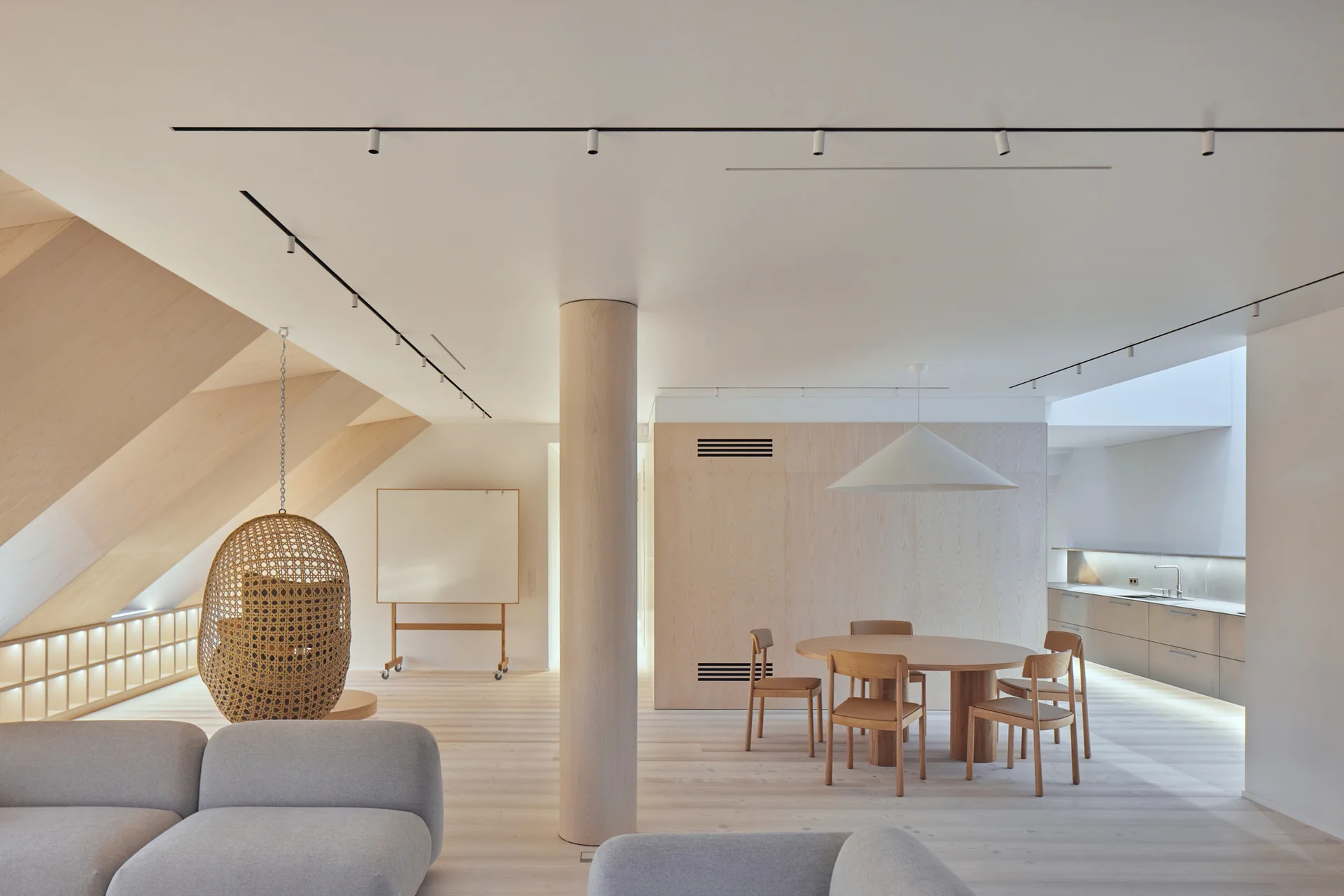 A minimalist open-plan living and dining room with a central round column, sloped wood ceiling, woven hanging chair, and round dining table.
