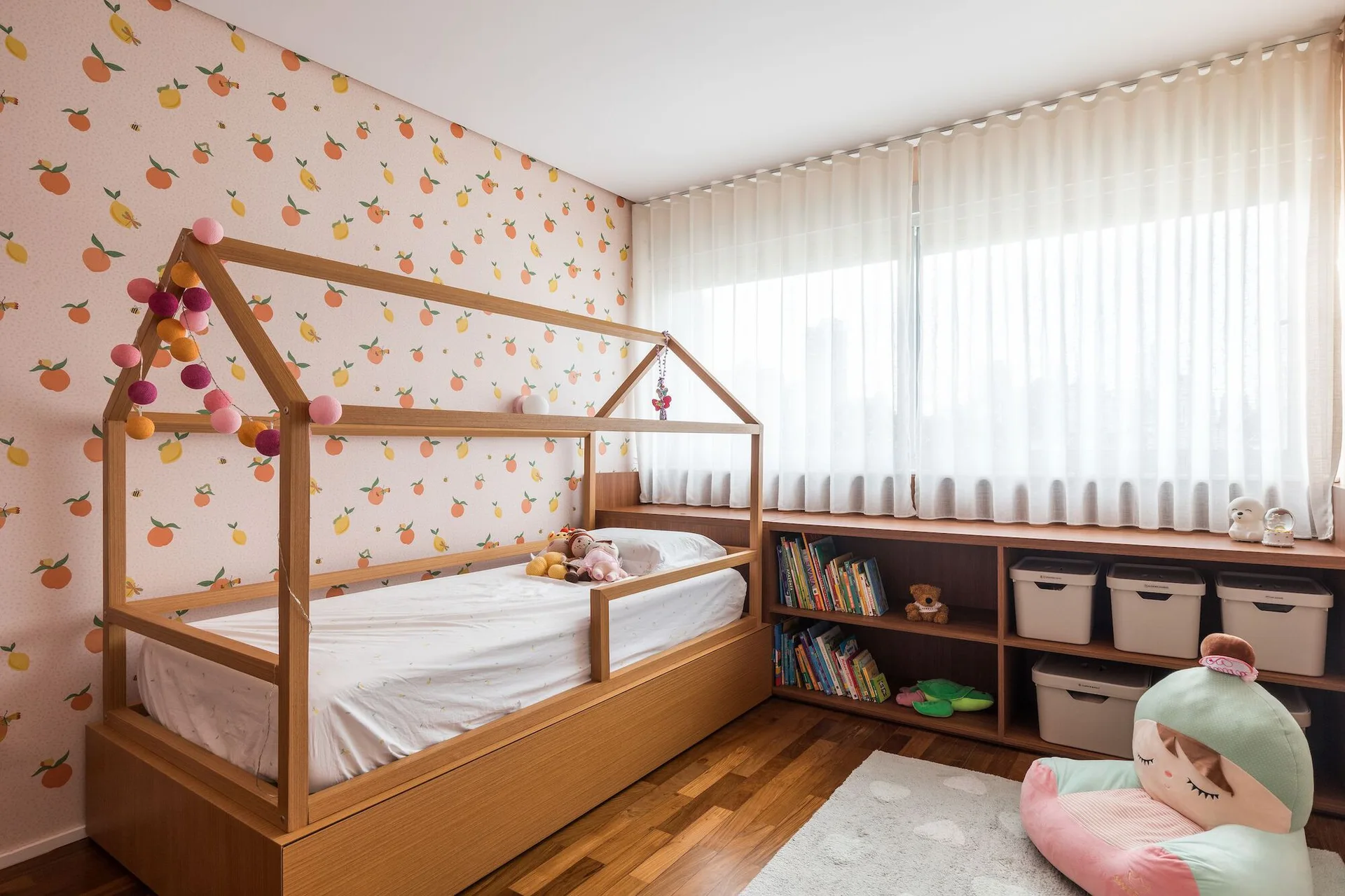 A children's bedroom features a wooden house-frame bed, peach fruit-patterned wallpaper, and a low wooden bookshelf under sheer white curtains.