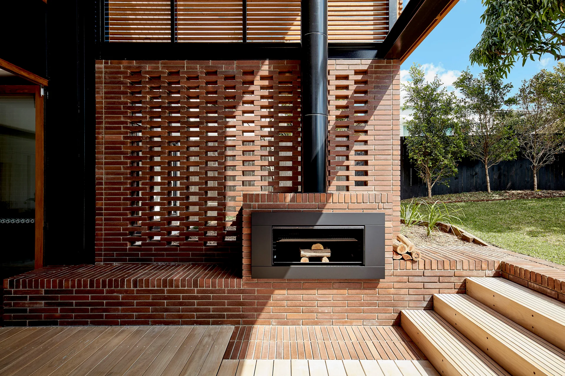An outdoor wood deck features a black metal fireplace integrated into a red brick wall with a perforated pattern, next to wooden steps.