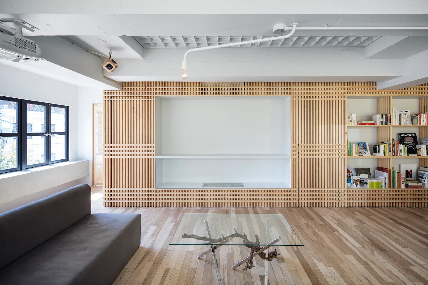 A minimalist living room features a light wood lattice wall unit, a grey sofa, and a glass-top coffee table on light wood floors.