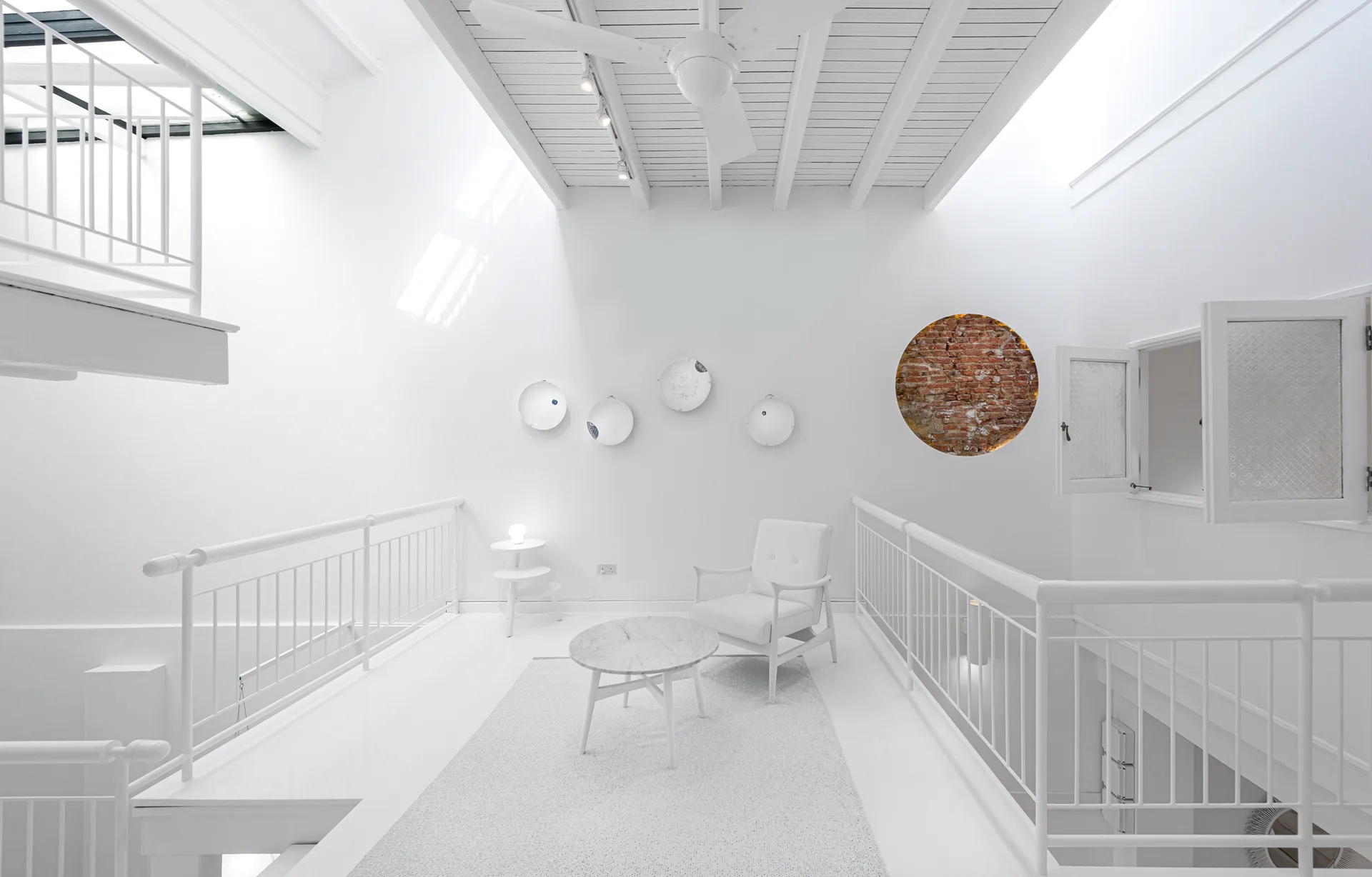 A bright white loft landing featuring a modern armchair, a round marble coffee table, white railings, and a circular wall cutout displaying red brick.
