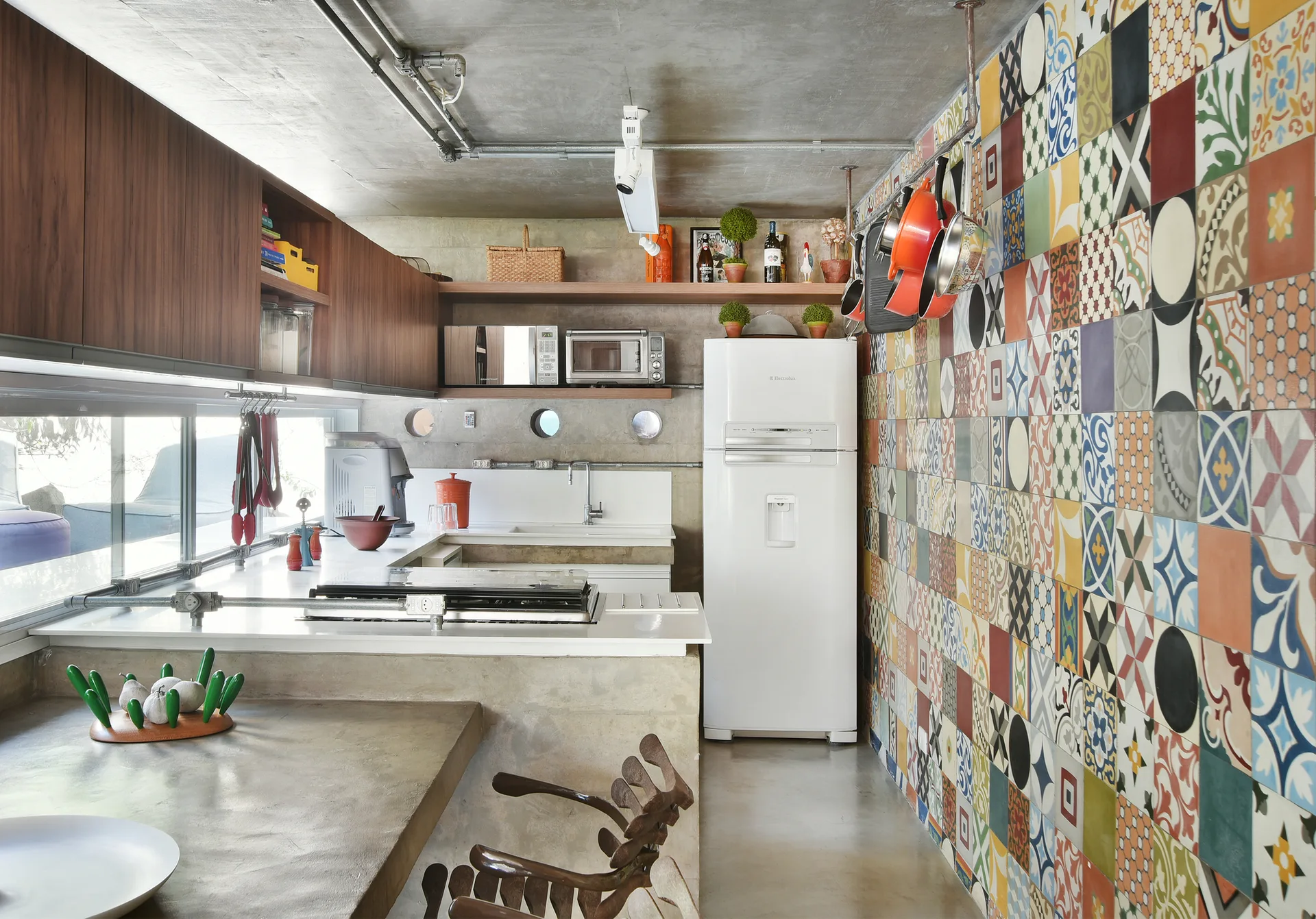 An industrial kitchen featuring a multicolored patterned tile accent wall, raw concrete ceiling and countertops, wood cabinets, and a white refrigerator.