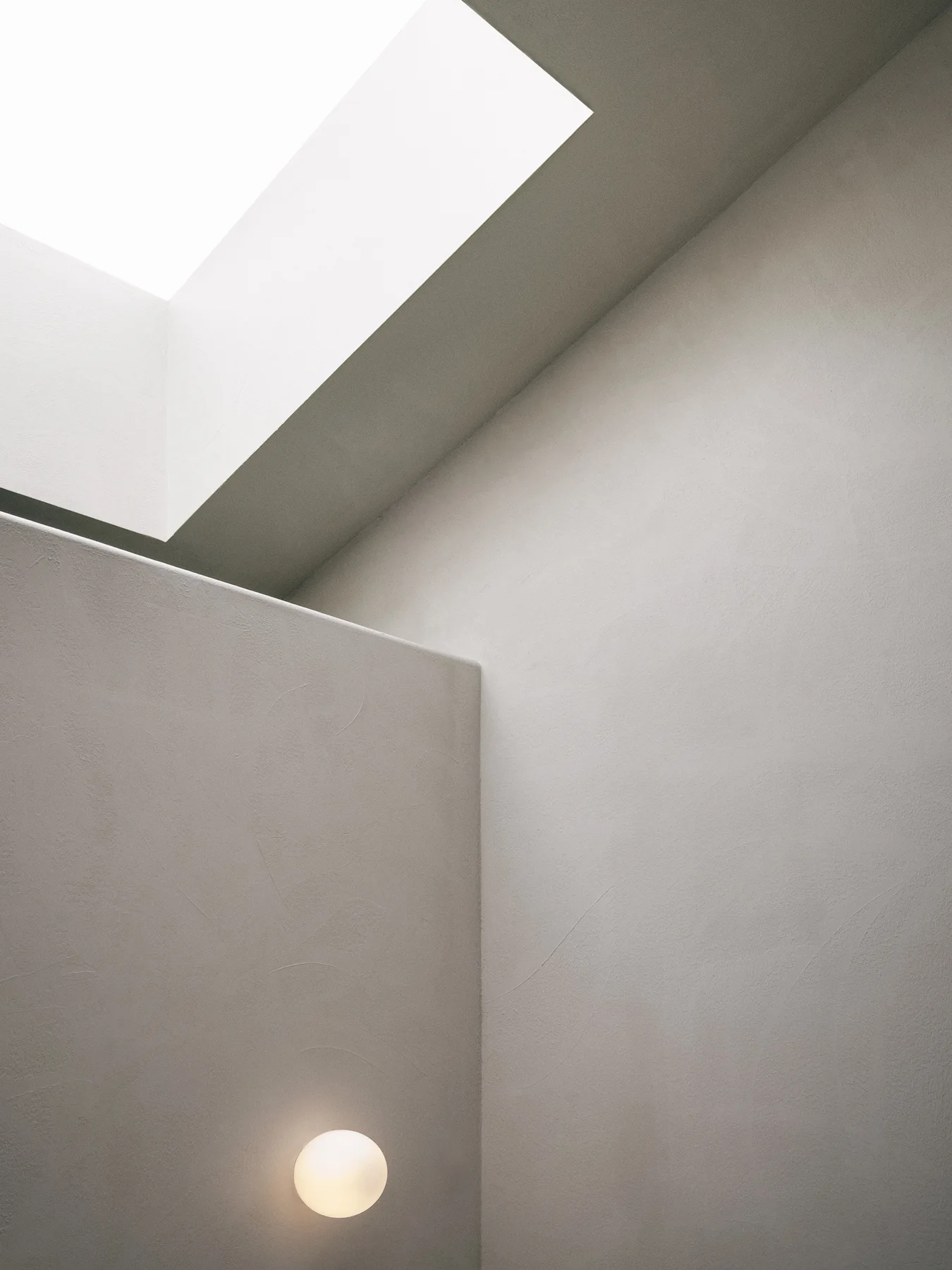 A minimalist architectural corner features textured greige plaster walls, a bright recessed skylight, and a glowing circular wall light.