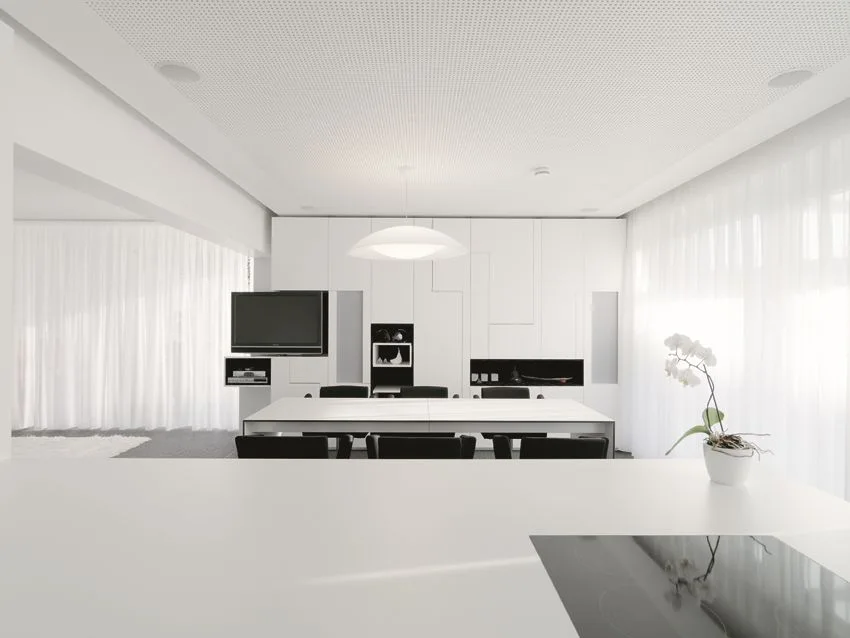 A minimalist dining room features a white table with black chairs beneath a pendant light and custom white wall cabinetry.