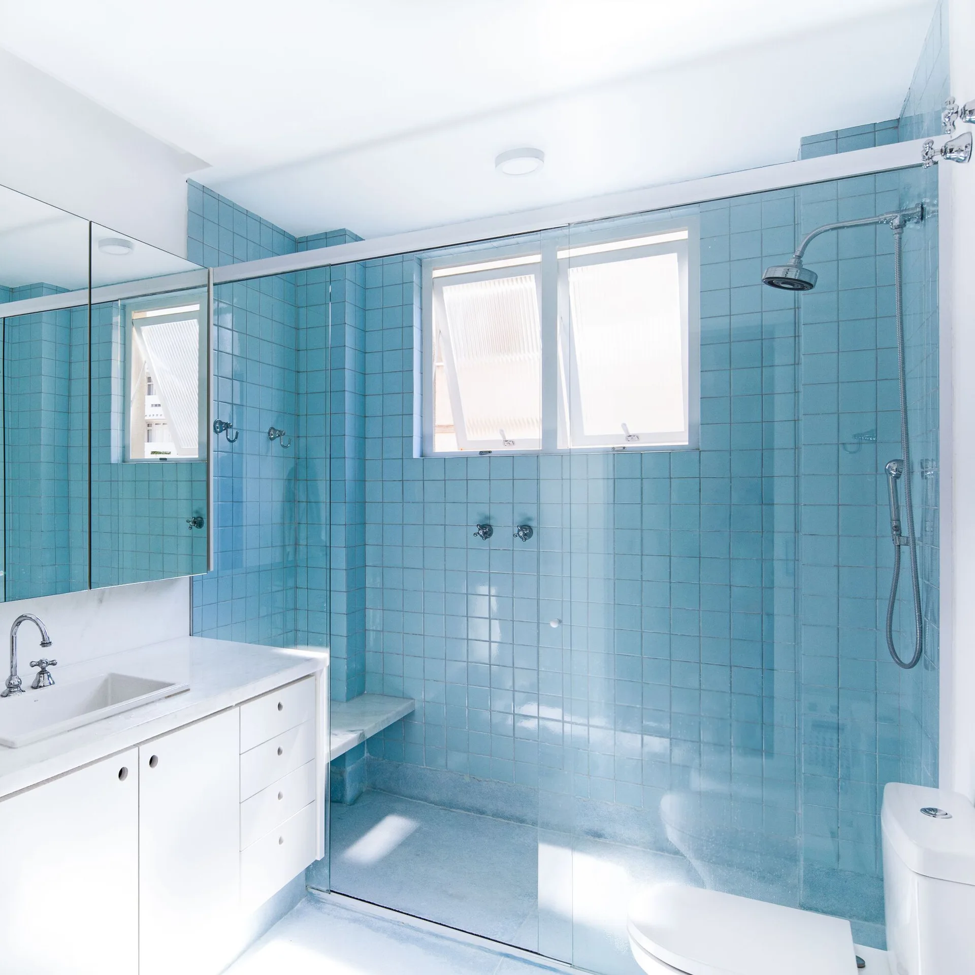 A modern bathroom featuring a walk-in glass shower lined with light blue square tiles, a white vanity with a sink, and a white toilet.