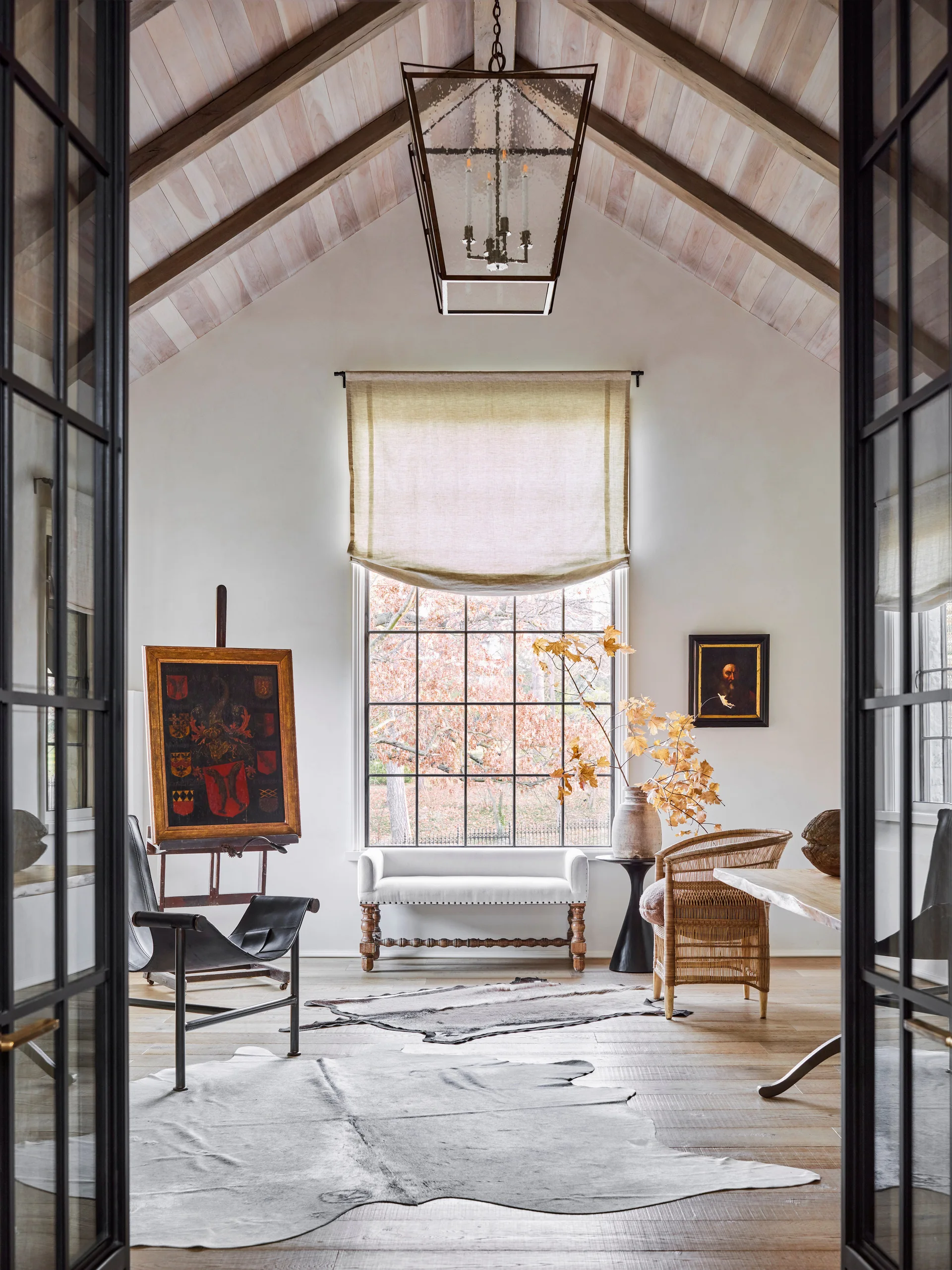 A study viewed through double glass doors features a vaulted wood ceiling, a large grid window, and a historic painting on an easel.