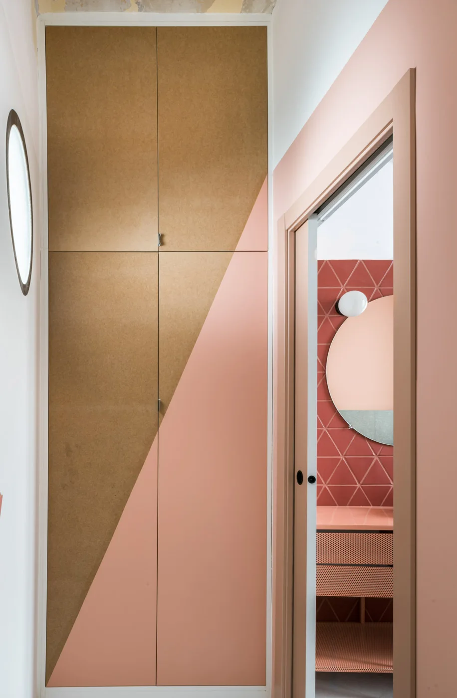 A tall hallway cabinet featuring diagonal pink color blocking stands next to a doorway looking into a bathroom with geometric red wall tiles.