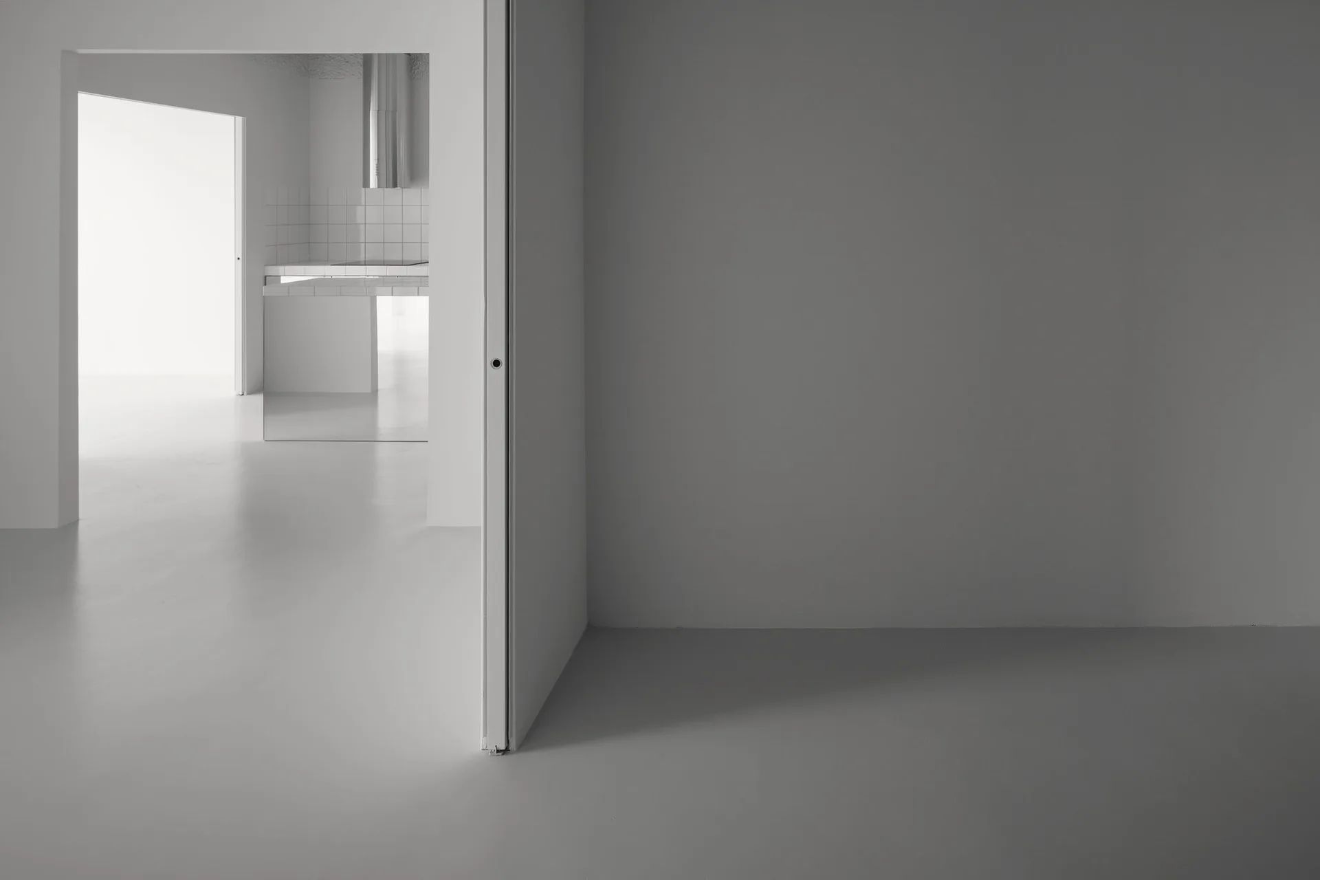 A minimalist home interior with solid gray walls and floors leading through an open doorway to a kitchen with white grid tiles.