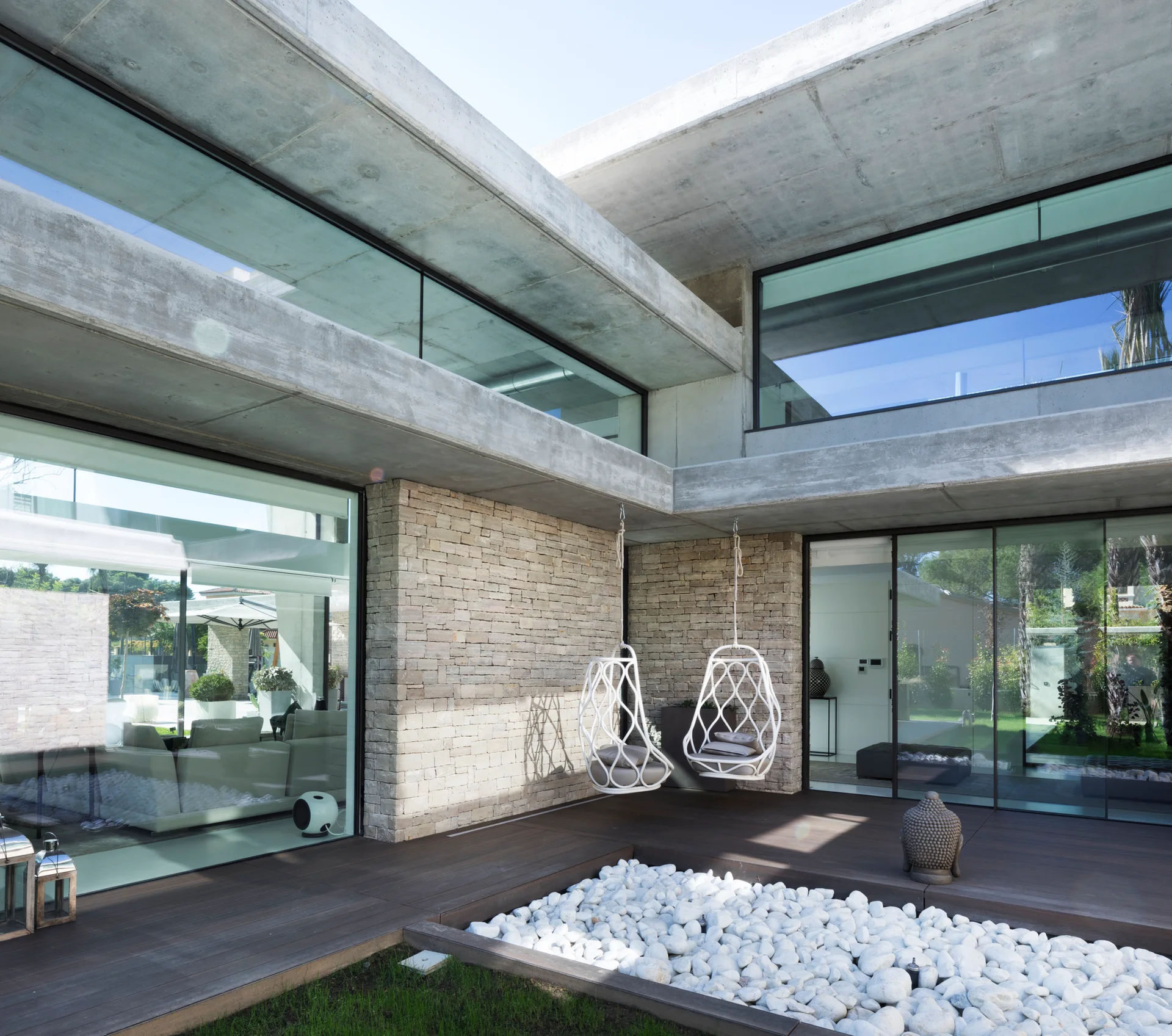 A modern outdoor patio featuring a dark wood deck, concrete overhangs, natural stone walls, two white hanging chairs, and a bed of white stones.