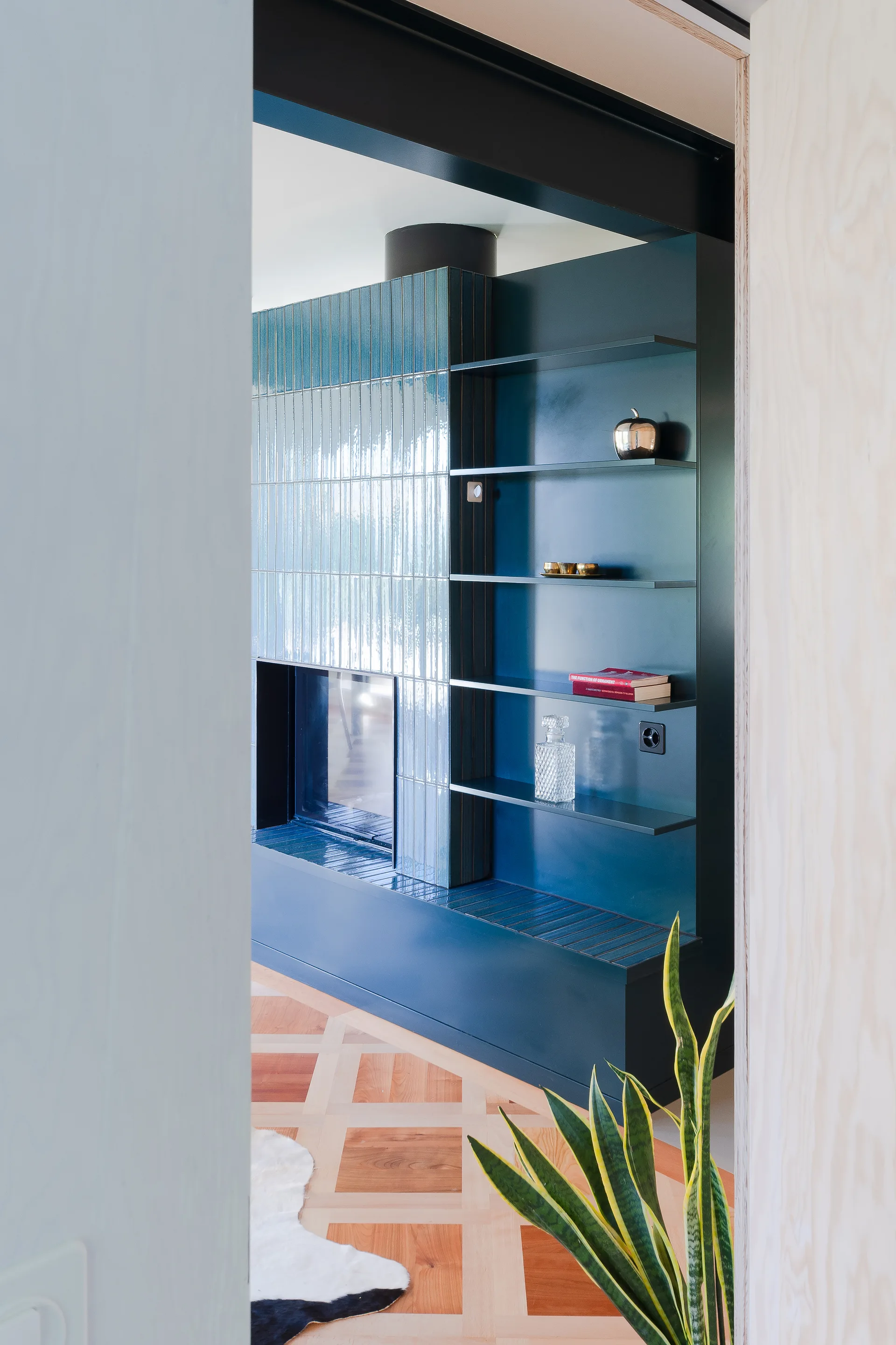 A view through a doorway shows a glossy blue tiled fireplace integrated with matte blue open shelving on a geometric parquet wood floor.