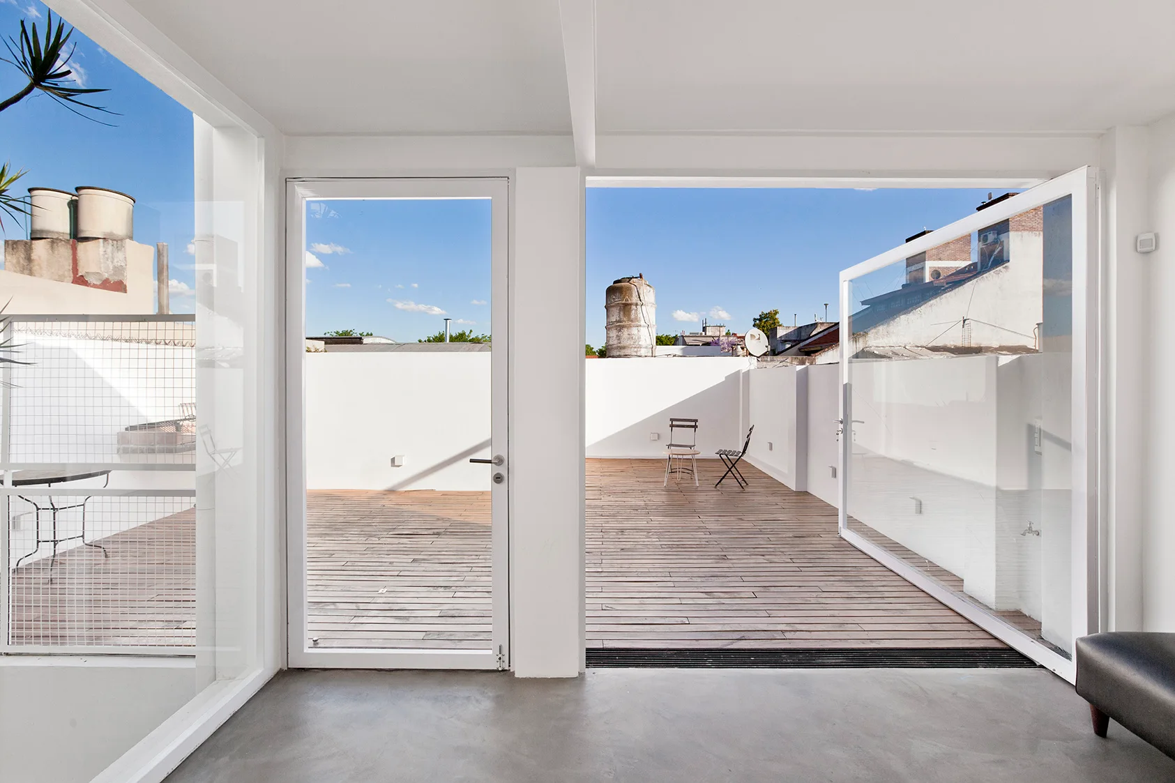 An interior space opens through swinging white-framed glass doors onto a rooftop terrace with wood plank decking and a clean white perimeter wall.