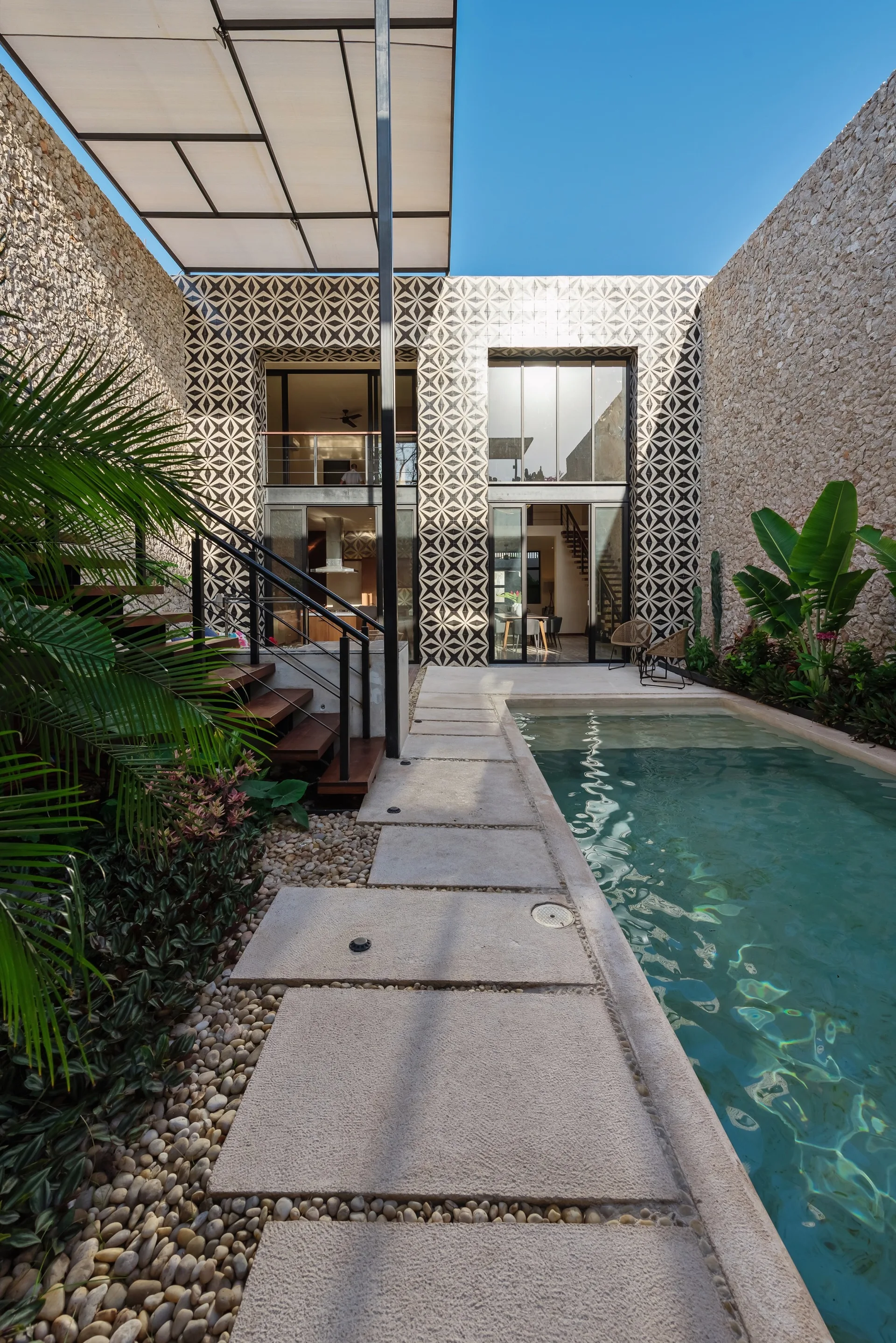 A modern outdoor courtyard featuring a rectangular swimming pool, concrete paver path over pebbles, rough stone walls, and a geometric tiled facade.