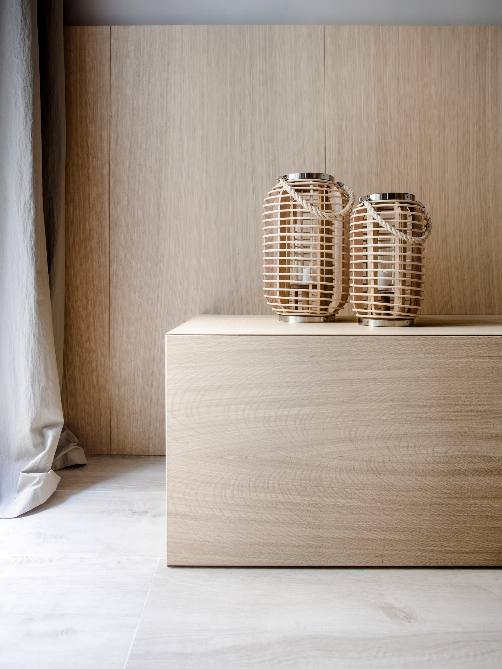 Two slatted wooden lanterns sit on a light wood block console table against a matching wood paneled wall next to a grey curtain.