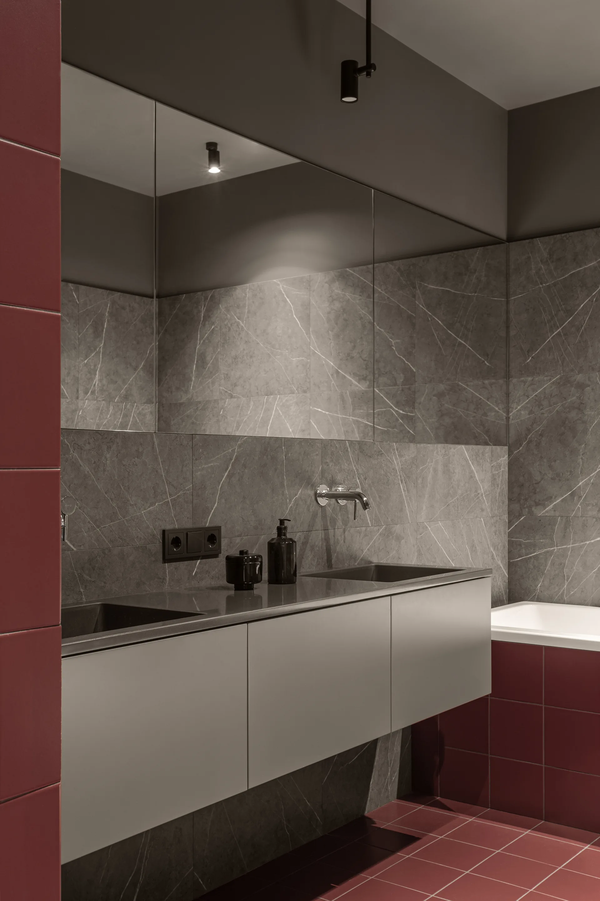 A contemporary bathroom features a matte grey floating vanity with a wall-mounted faucet, surrounded by veined stone walls and deep red floor tiles.
