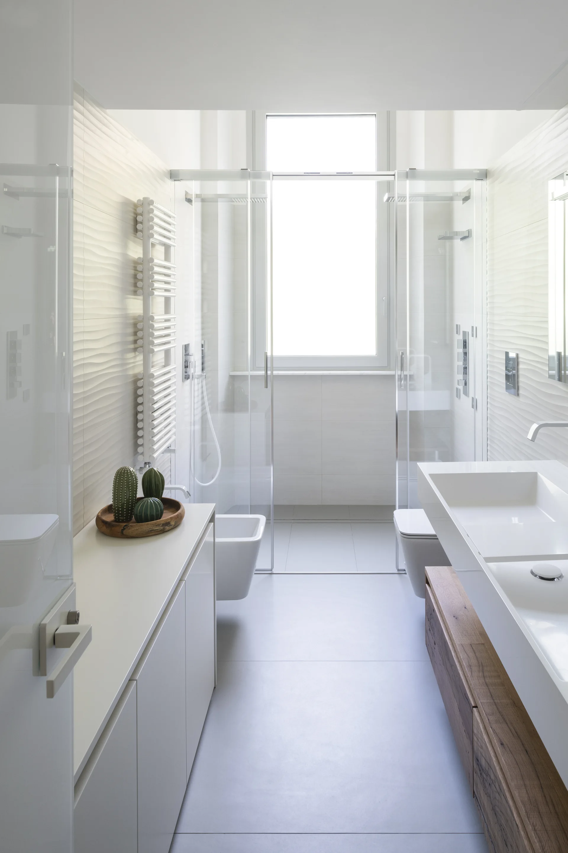 A narrow modern bathroom featuring textured wave-patterned wall tiles, a floating wooden vanity with white sinks, and a glass-enclosed walk-in shower.