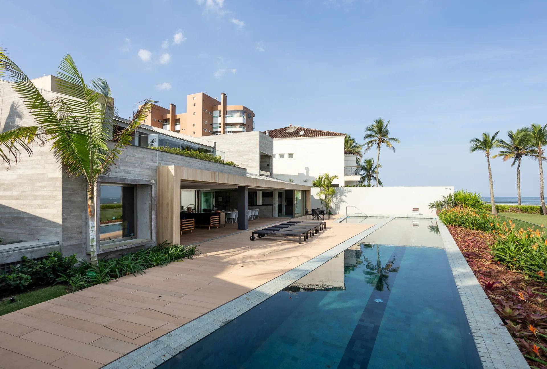 A long rectangular swimming pool with blue water next to a modern concrete house with warm wood-toned plank decking, black loungers, and palm trees.