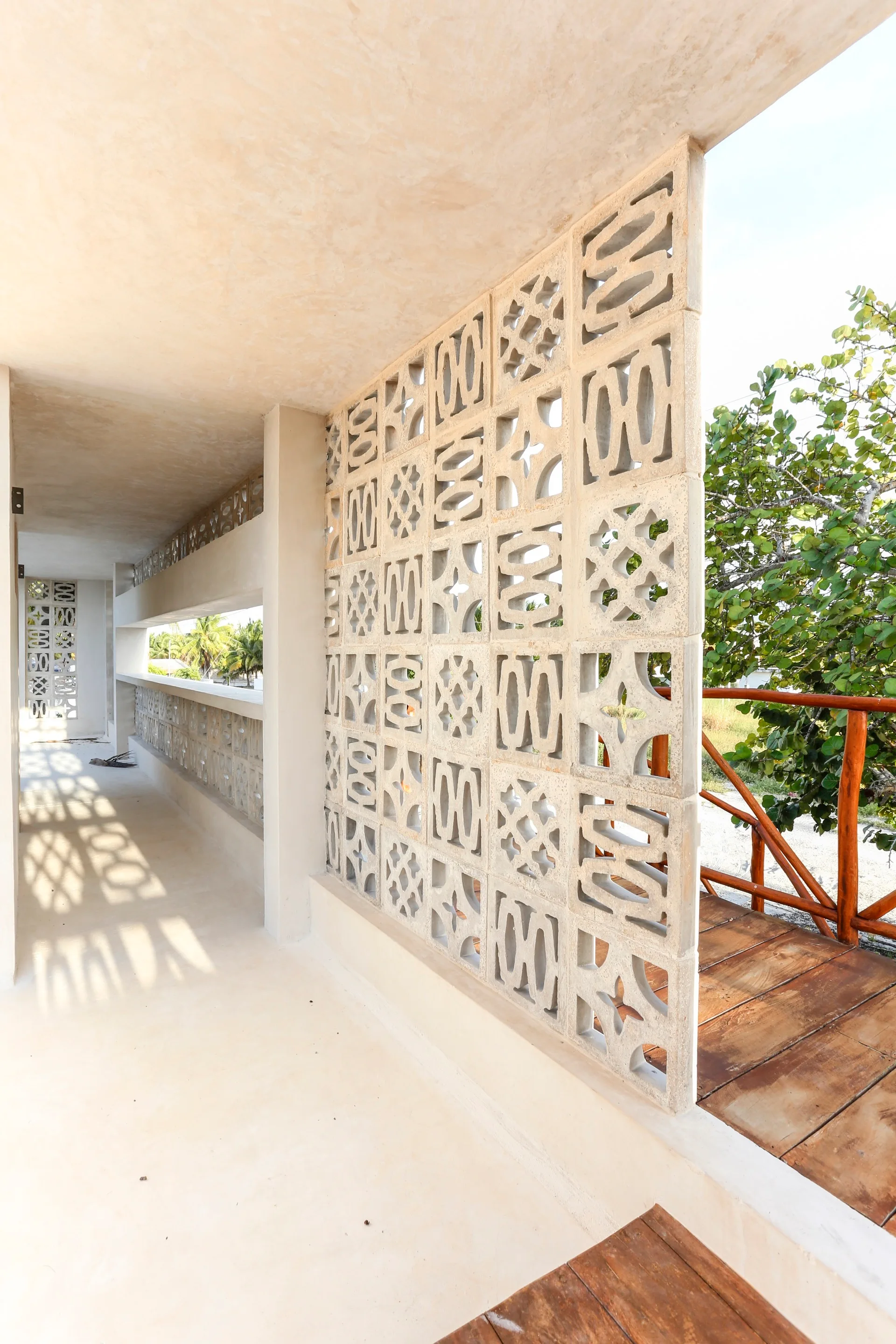 A covered outdoor corridor featuring a patterned concrete breeze block screen wall, stucco surfaces, and an adjacent wooden deck with a simple railing.