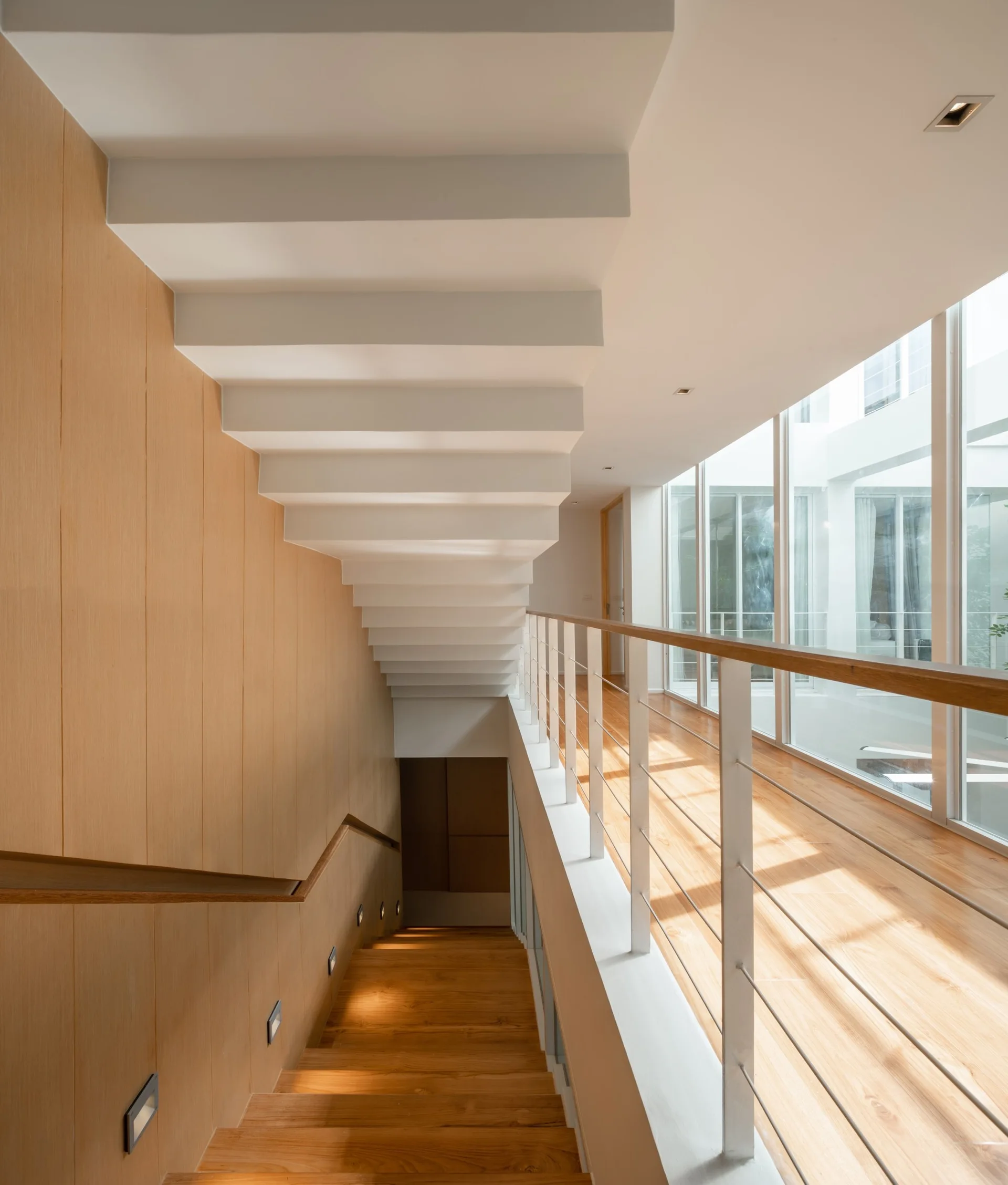 A modern staircase with wood-paneled walls, wooden steps, and a white undersided upper flight runs alongside a glass hallway with a cable railing.