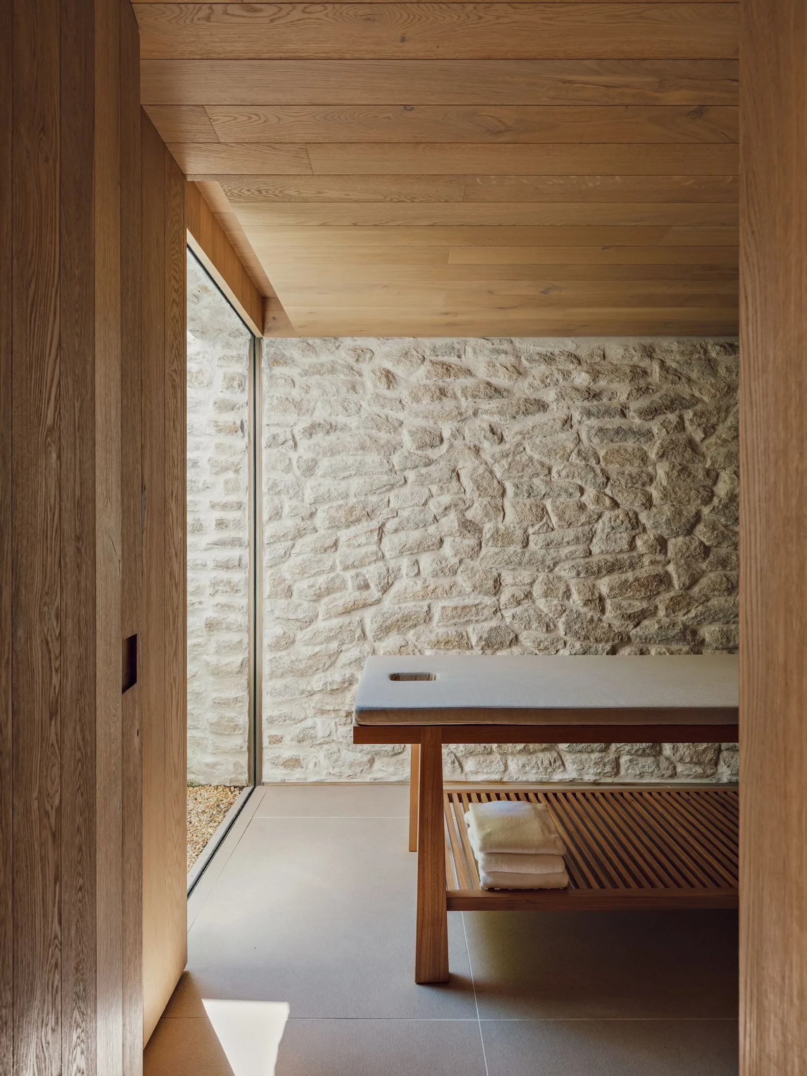 A wooden massage table stands in a wellness room featuring a rough stacked stone wall, a wood-paneled ceiling, and tiled floors.
