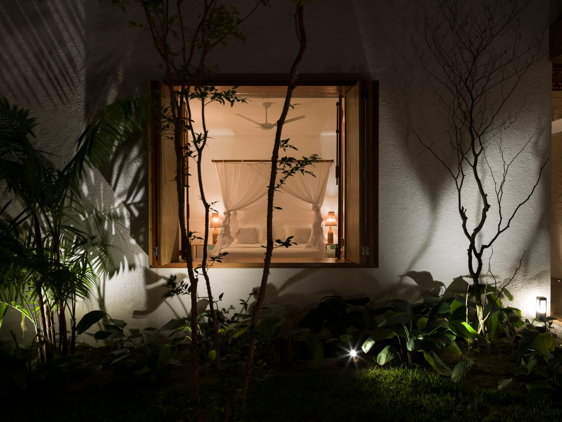 A view through an open wooden window frame into a brightly lit bedroom with a white canopy bed and glowing bedside table lamps.