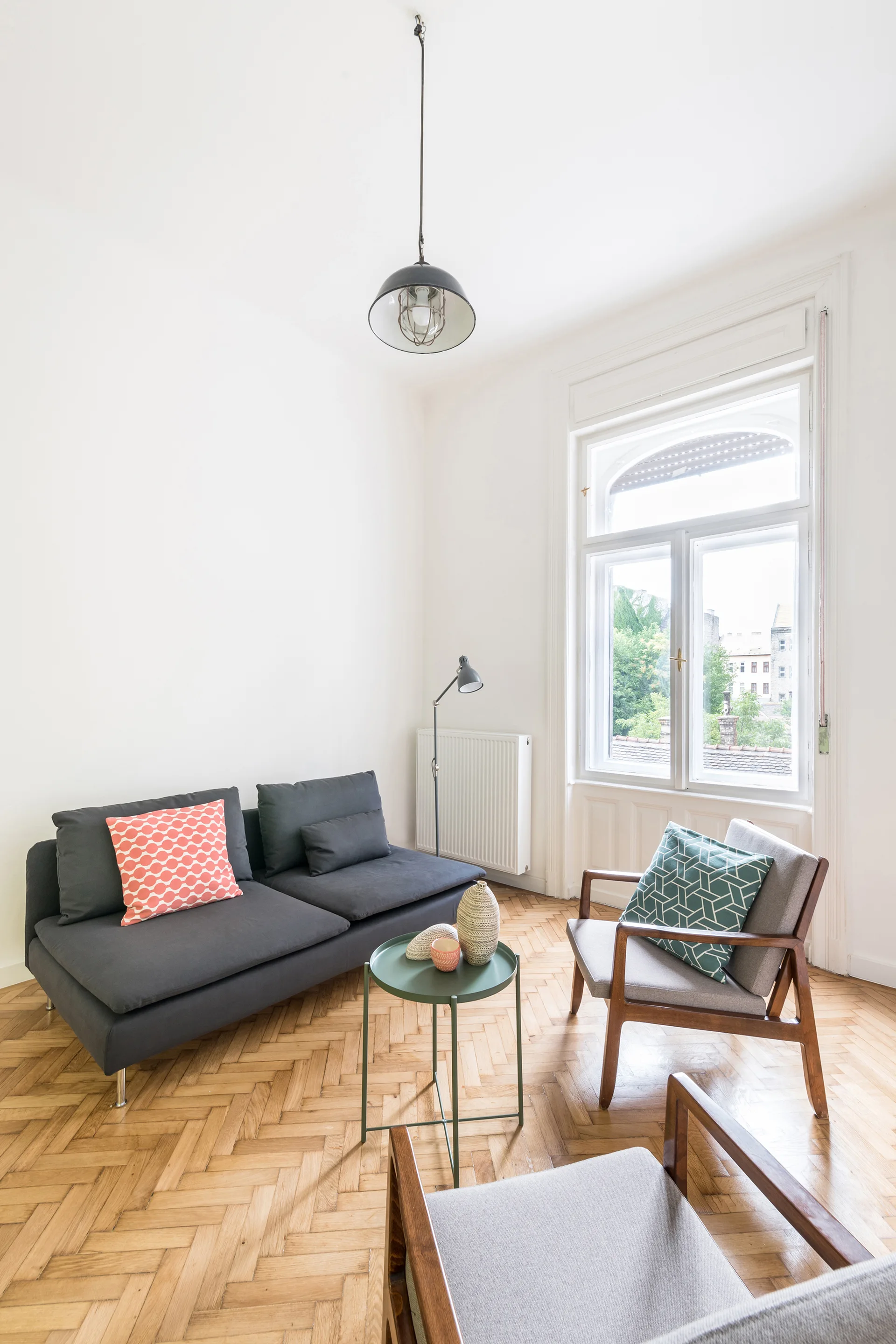 A bright living room features a dark grey sofa, two wooden armchairs with pattern cushions, and a green coffee table on herringbone parquet flooring.
