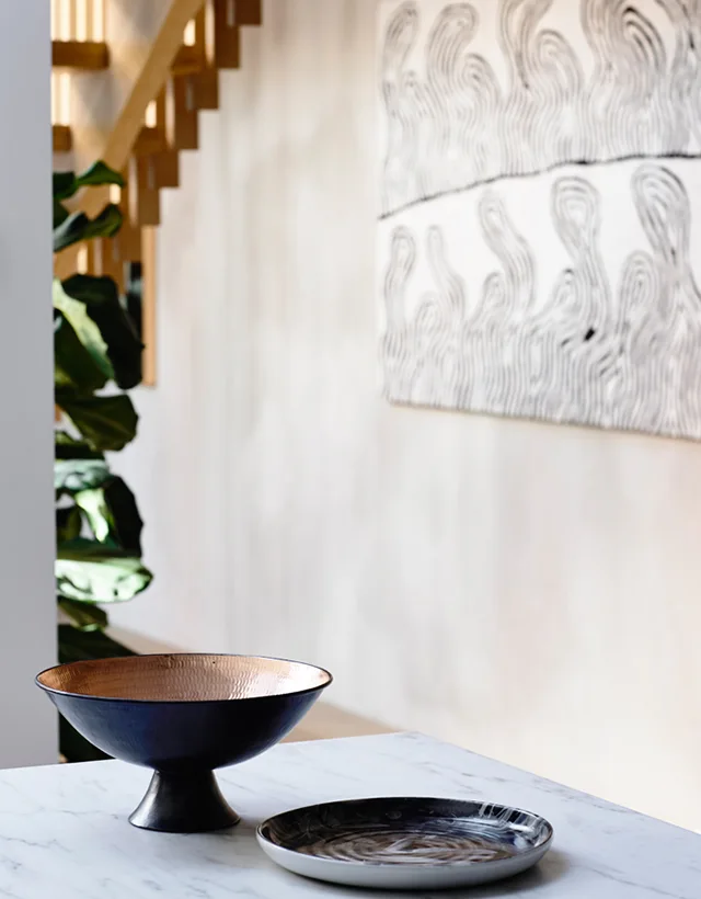 A black and copper pedestal bowl and a swirled decorative plate rest on a white marble tabletop in front of blurred stairs and wall art.