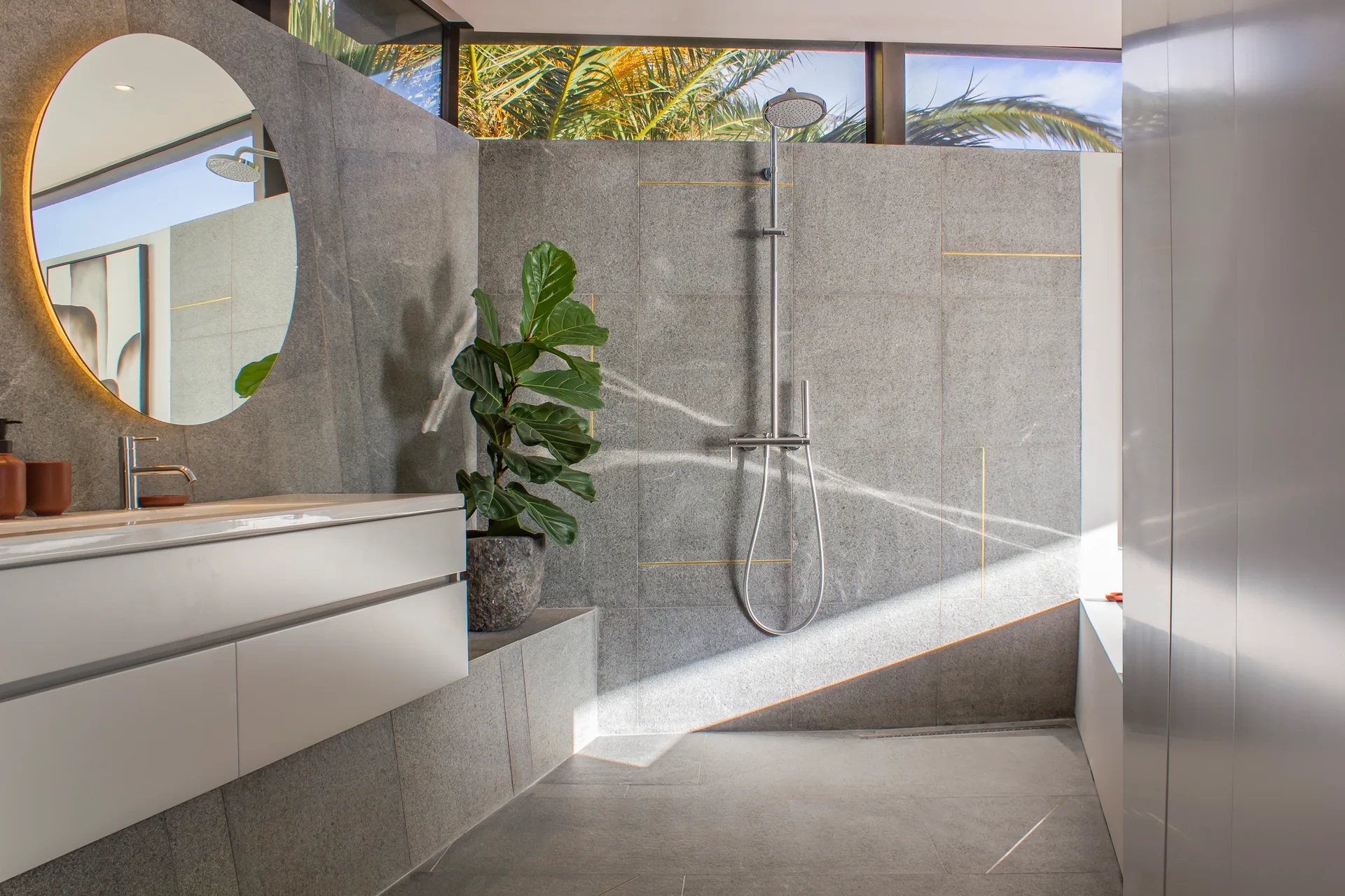 A modern grey tiled bathroom featuring a white floating vanity, a backlit circular mirror, a sleek shower fixture, and a clerestory window.