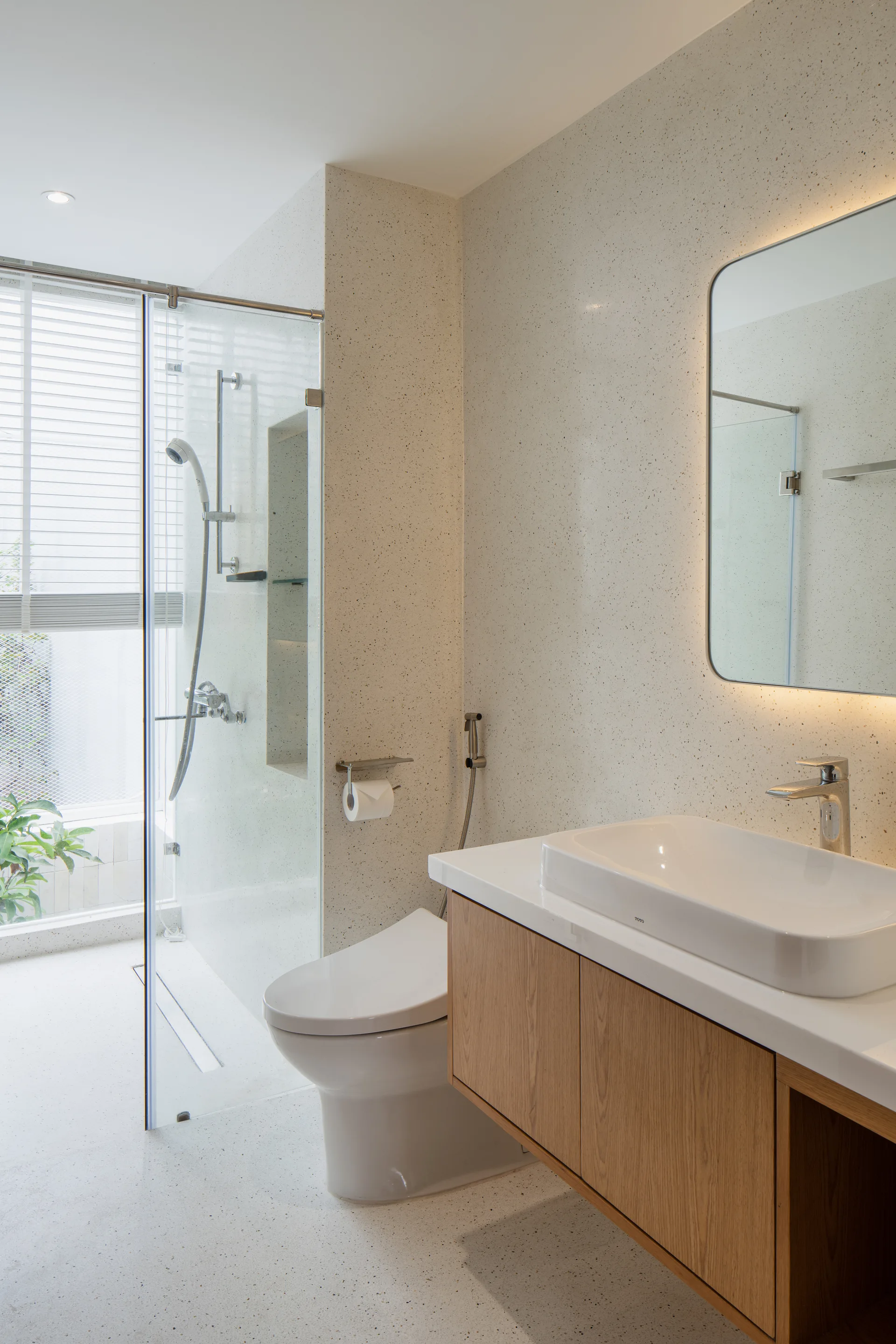 A modern bathroom features speckled terrazzo walls and floors, a wooden floating vanity with a white sink, and a glass-enclosed shower.