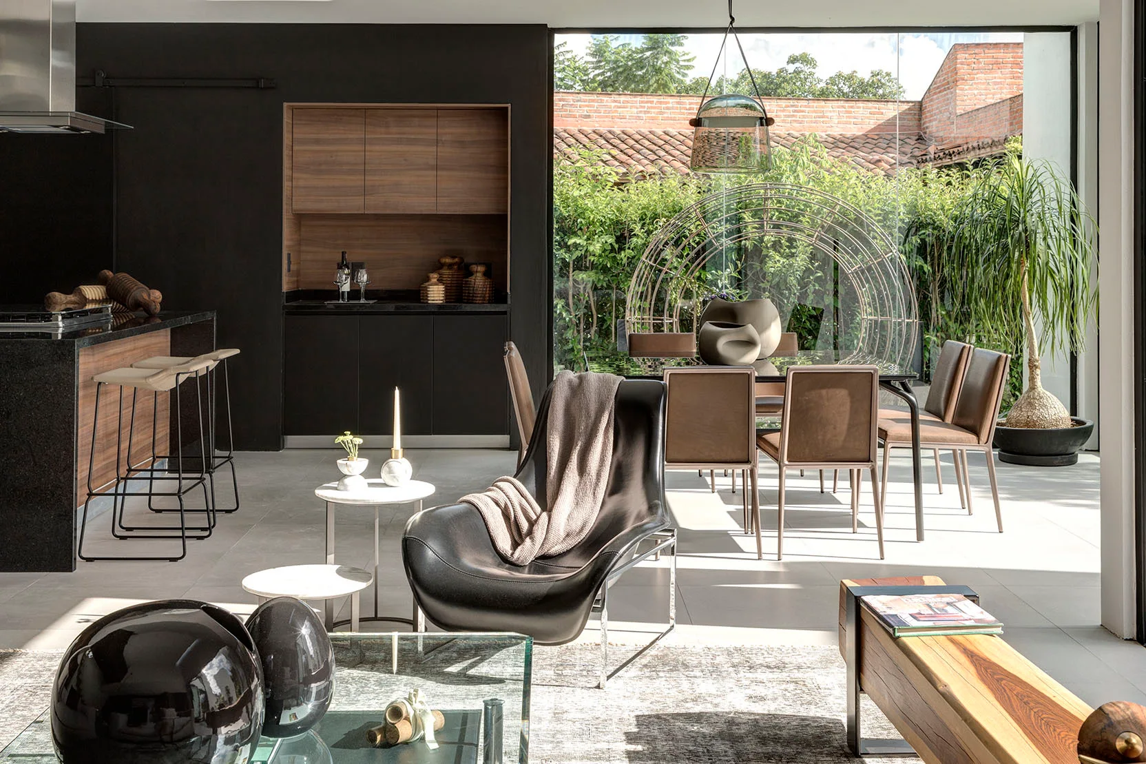An open-concept dining room features a glass-topped table with brown leather chairs, next to a matte black kitchen and a large window looking out onto a garden.