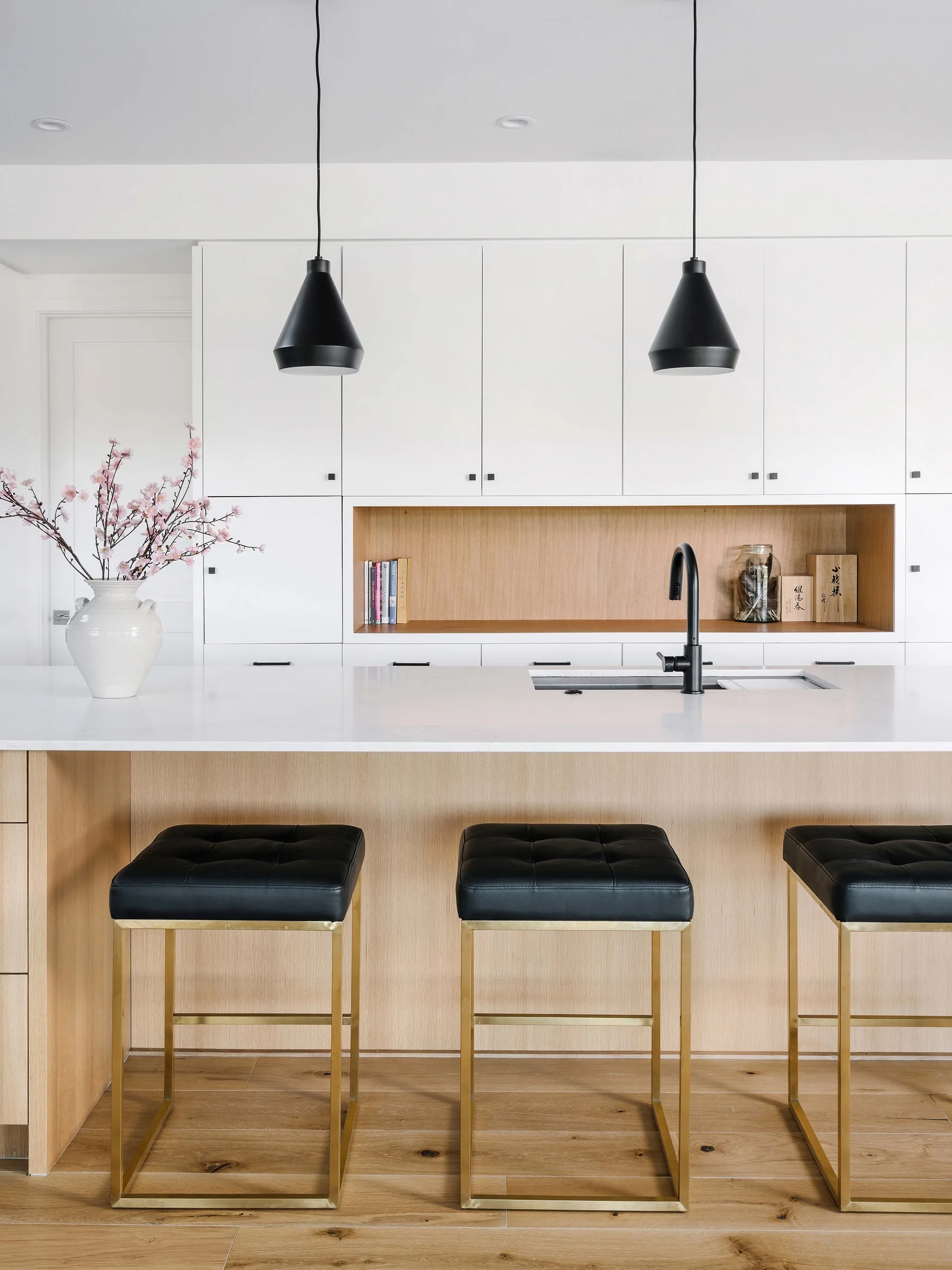 Three black tufted bar stools with brass frames sit at a white quartz kitchen island in front of minimalist white cabinets with a wood accent niche.