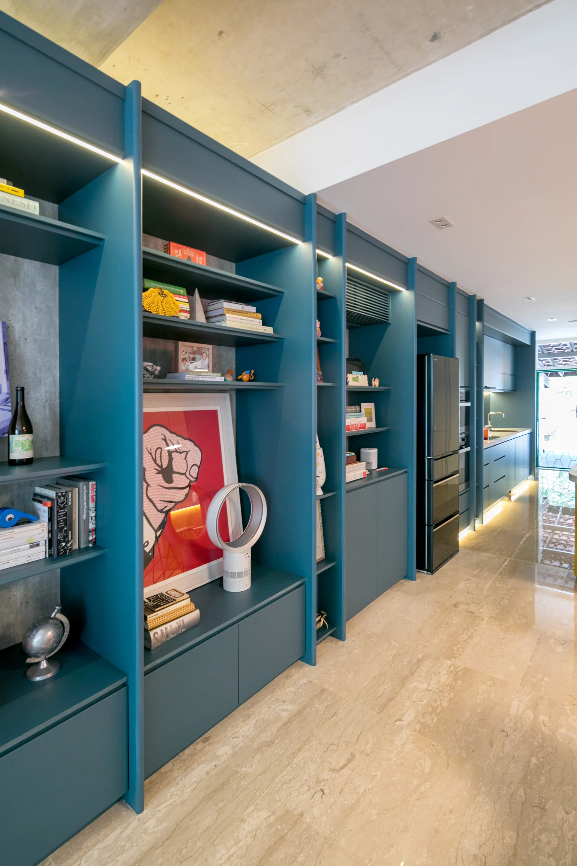 Teal built-in shelving and cabinets with LED strip lighting line a long kitchen hallway with a concrete ceiling and polished stone floor.