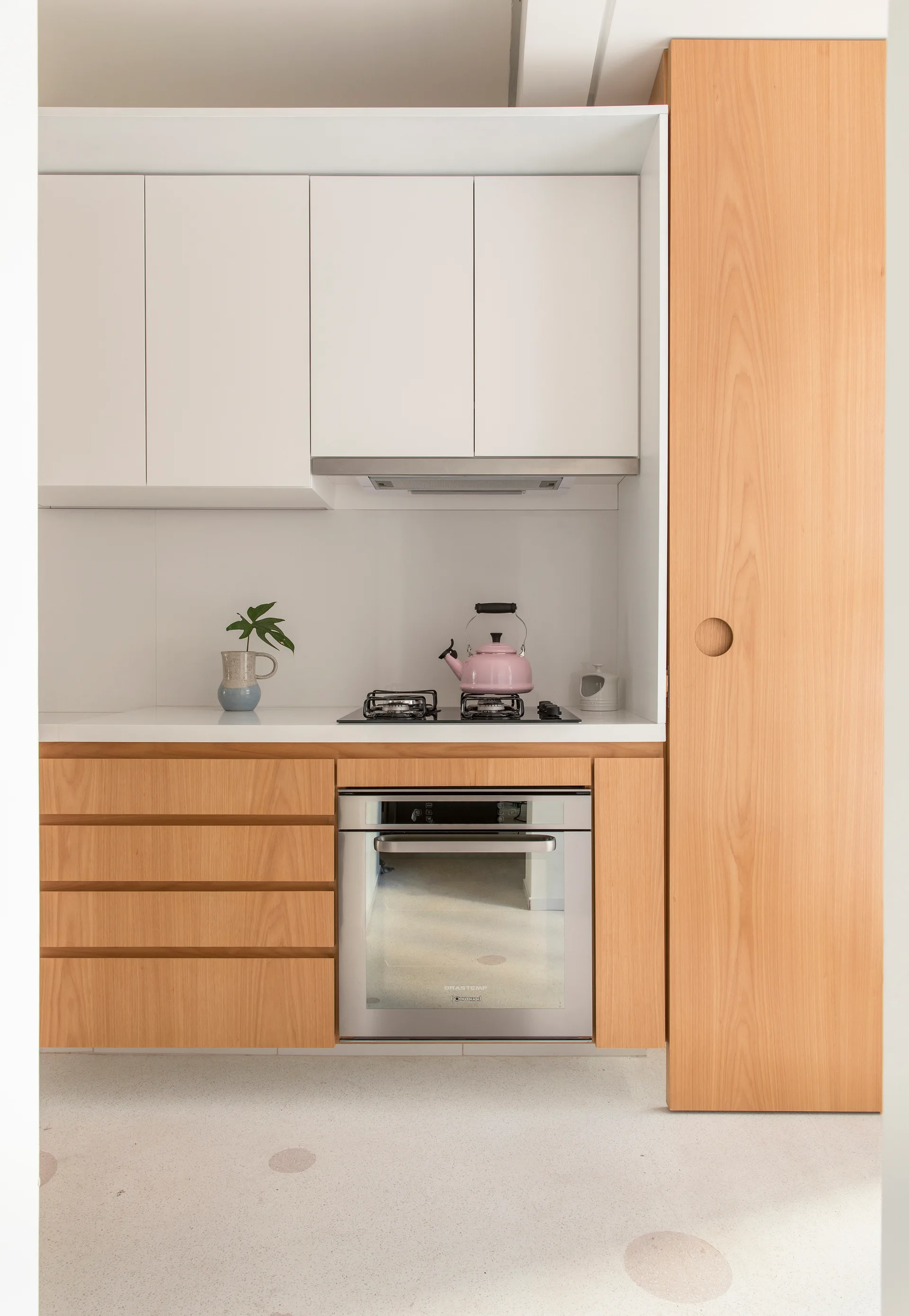 A minimalist kitchen features white upper cabinets, light wood lower drawers, a stainless steel oven, and a pink kettle on the gas cooktop.