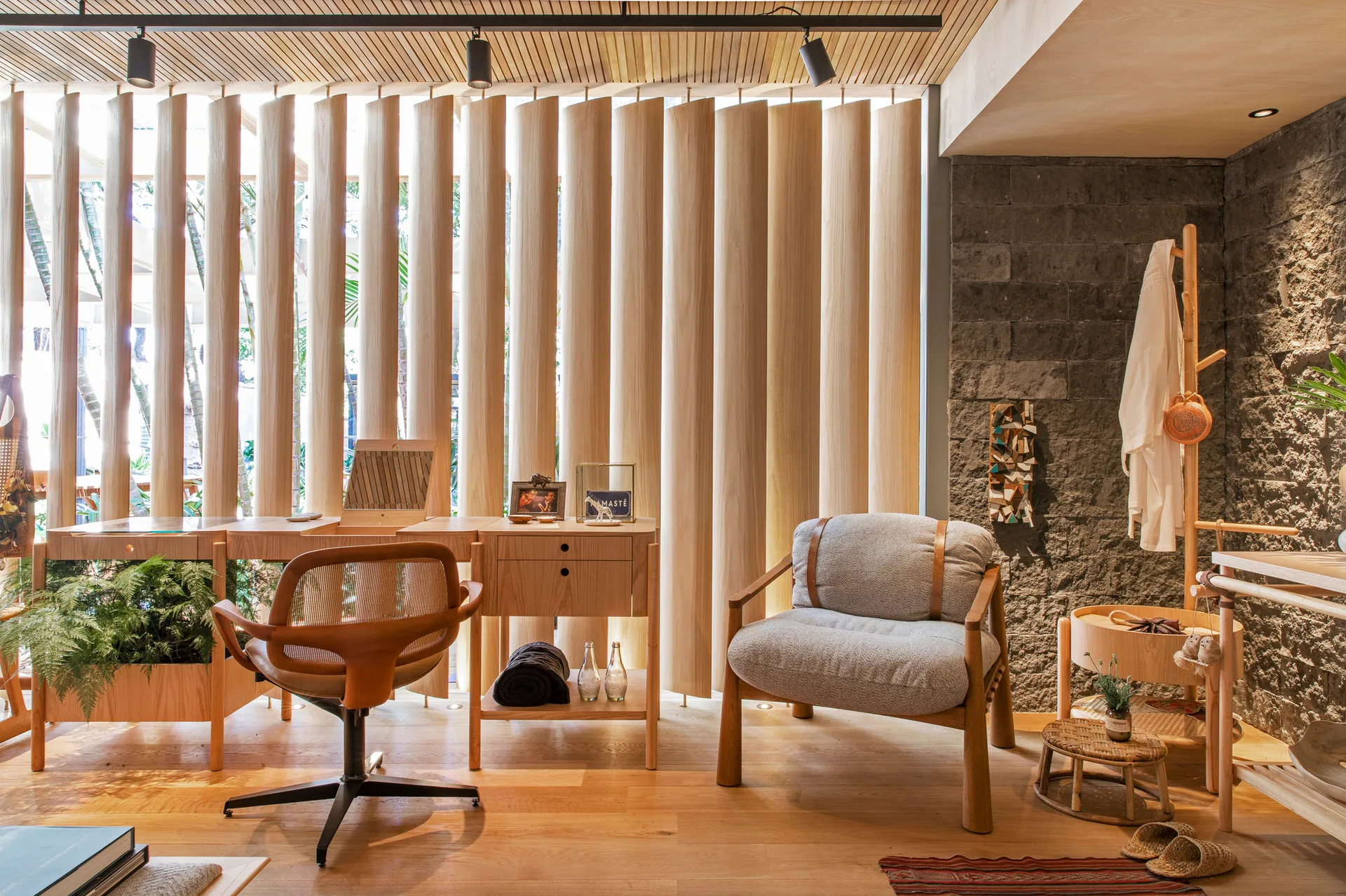A modern workspace featuring a light wood desk, a grey armchair, a vertical slatted wood divider, and a textured grey stone wall.