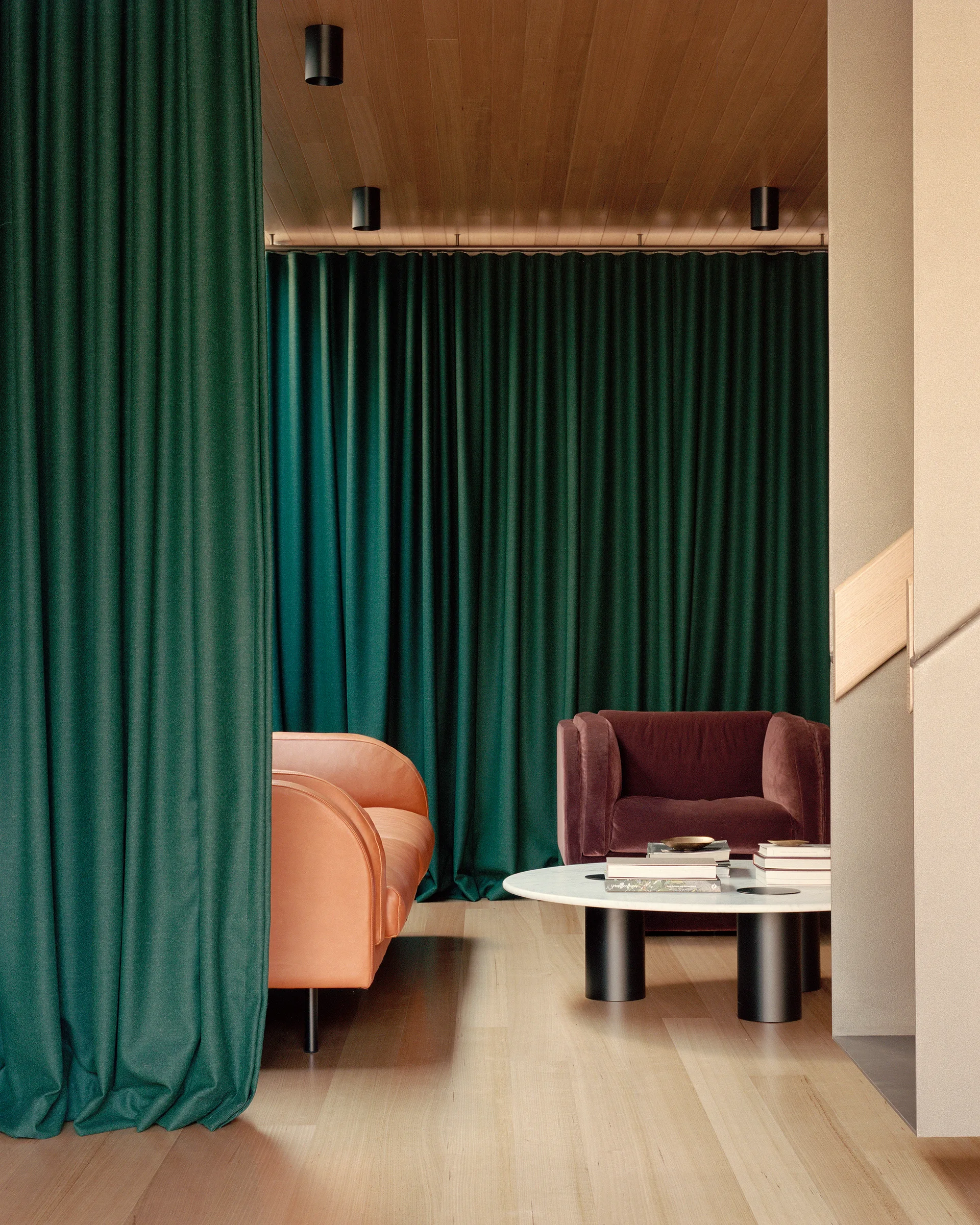 A contemporary lounge features an orange sofa and burgundy velvet armchair framed by a floor-to-ceiling green curtain and a wood-paneled ceiling.