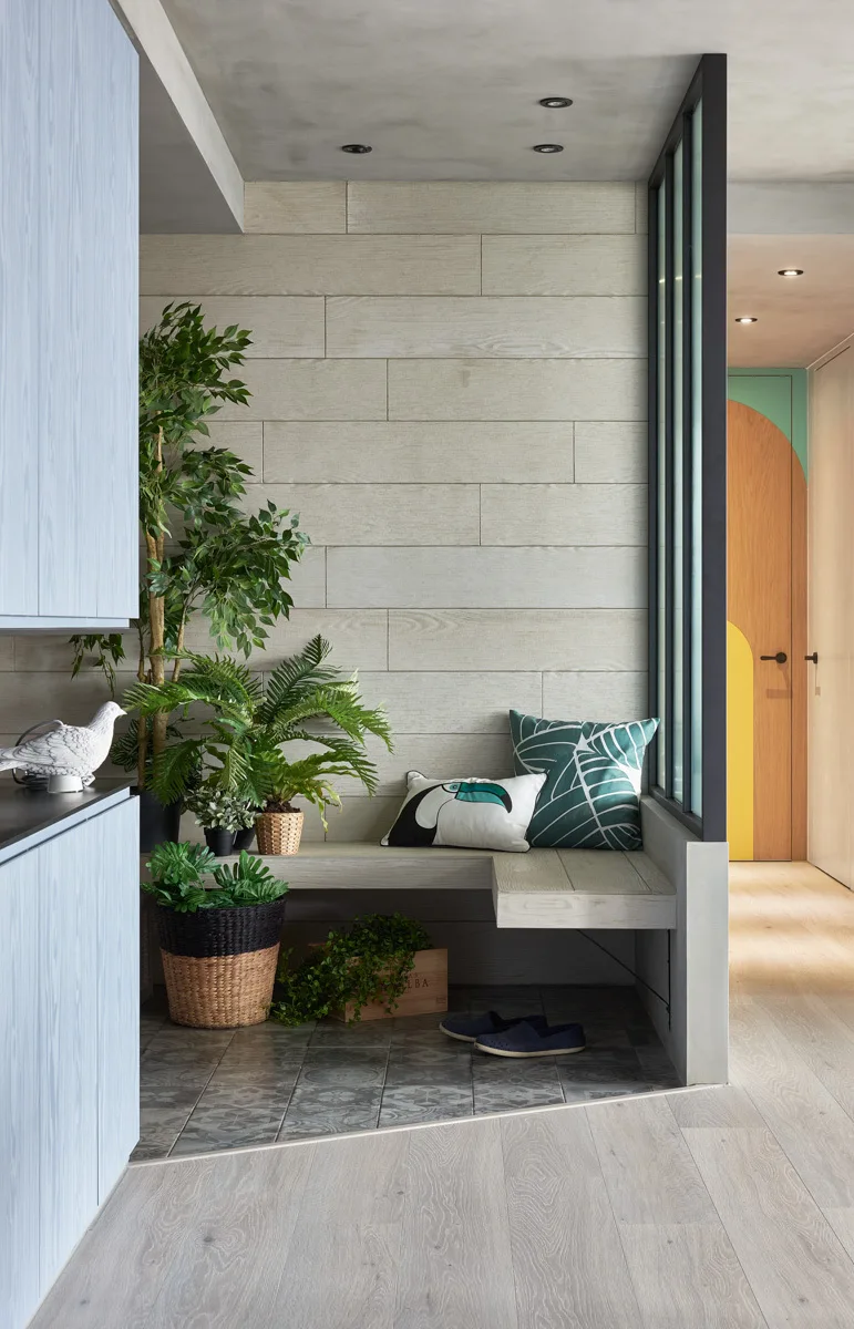 A modern entryway features an L-shaped built-in bench under a wood-paneled wall, set next to patterned floor tiles and a black-framed glass partition.