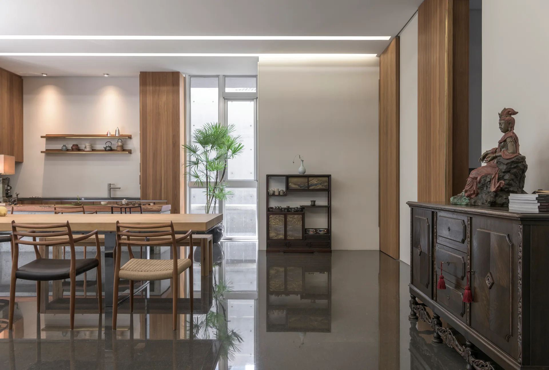 A minimalist dining room featuring a light wood dining table, woven chairs, a dark wood sideboard, and highly reflective dark floors.
