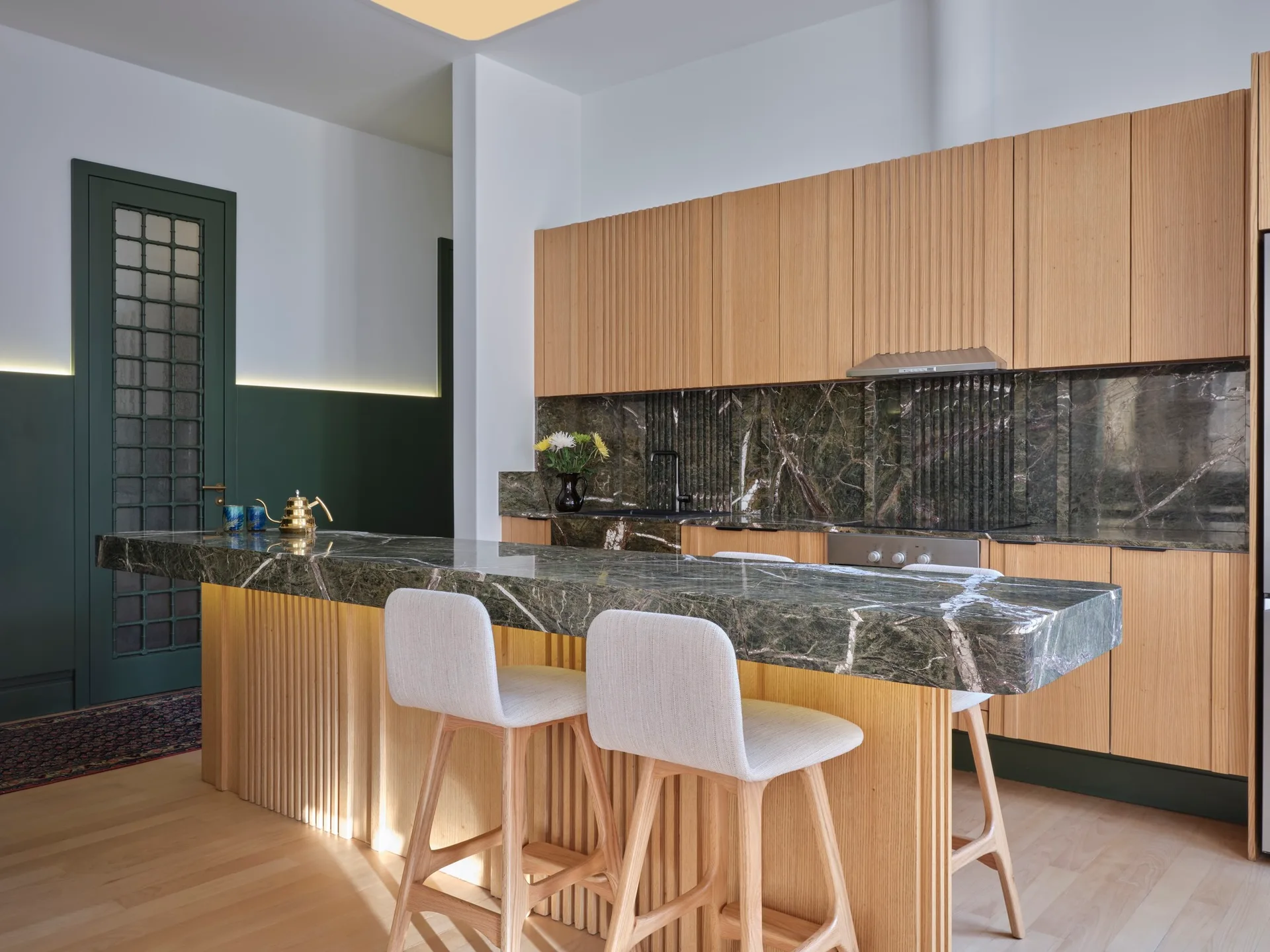 A modern kitchen featuring fluted oak cabinets, a dark green marble island with white bar stools, and a matching marble backsplash.