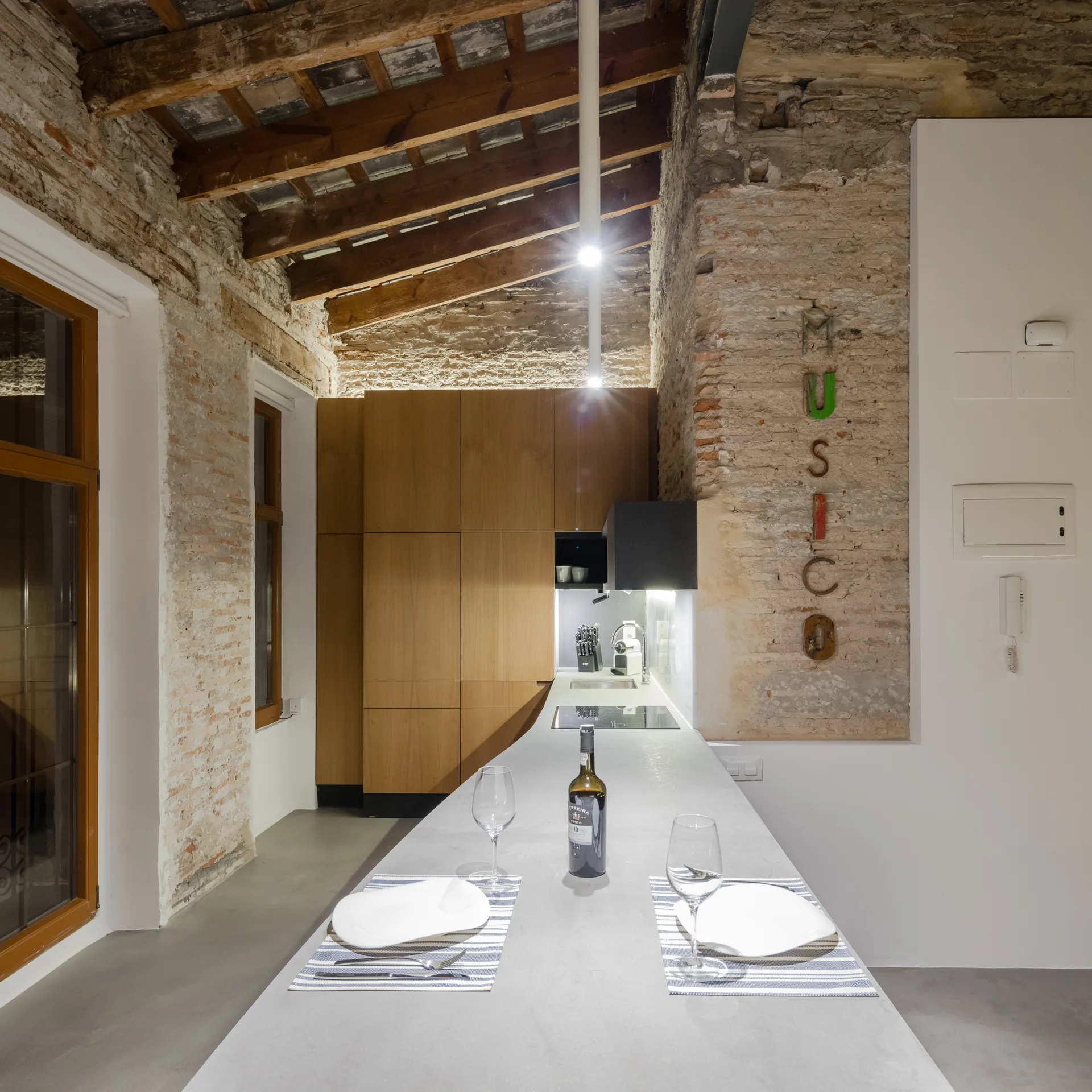 An industrial kitchen features a long concrete island set against rustic exposed brick walls and exposed wood ceiling beams.