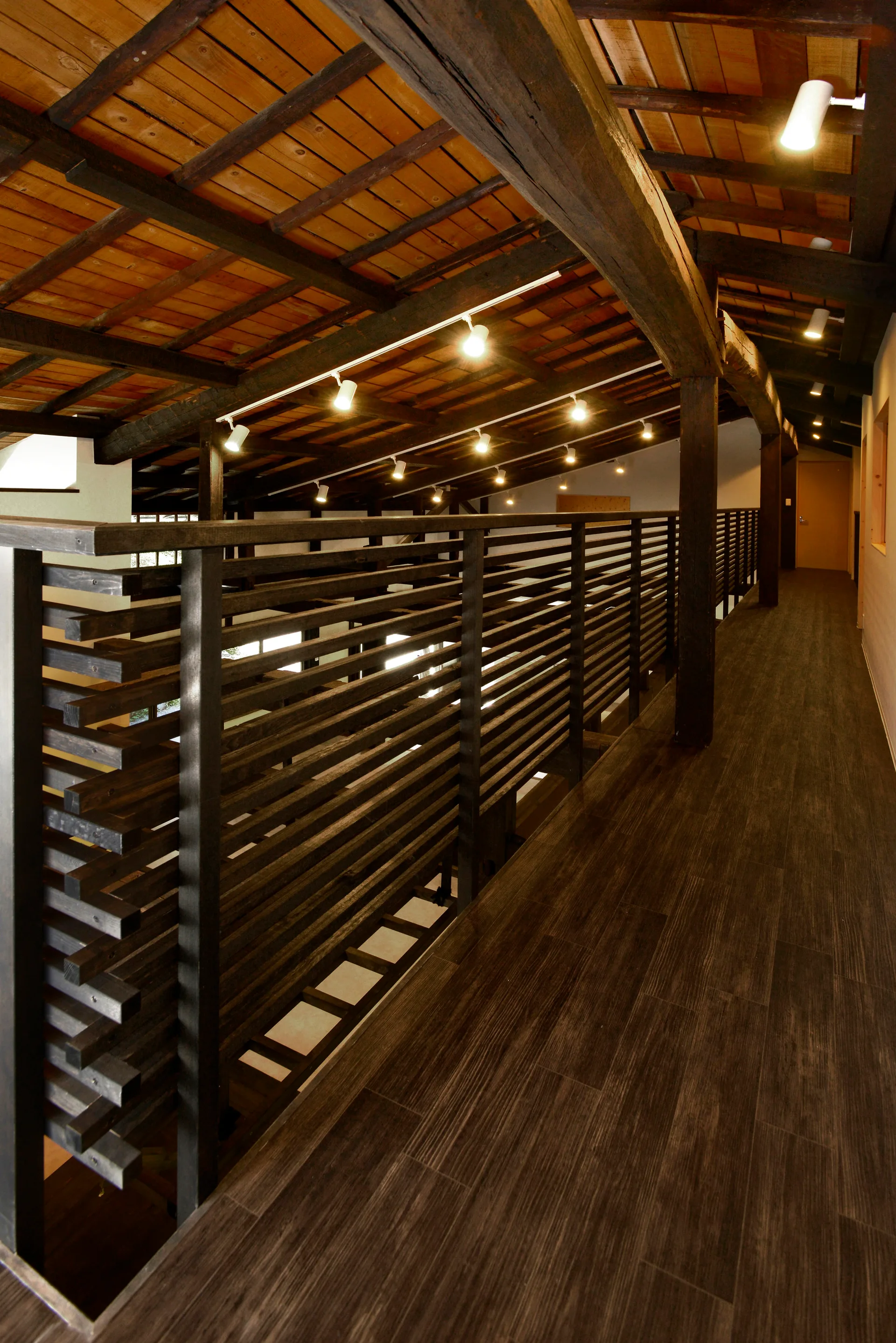 An elevated wood walkway features a dark slatted railing, wood-plank flooring, and an exposed timber ceiling with track lighting.
